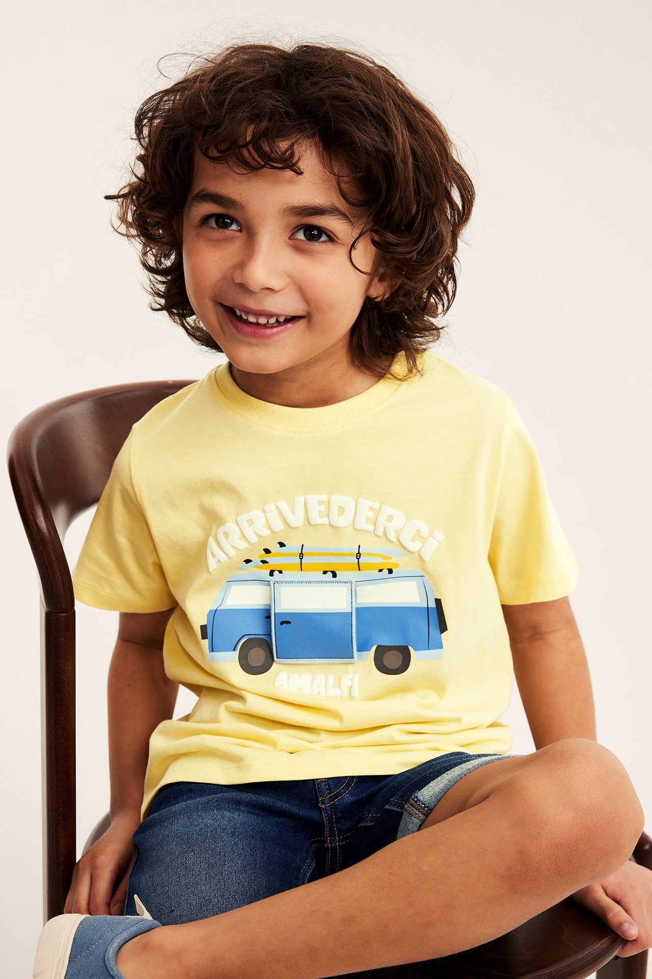 Framifrån: Leende barn i gul oversize bomulls-T-shirt med blå surfbilstryck och mörkblå shorts.