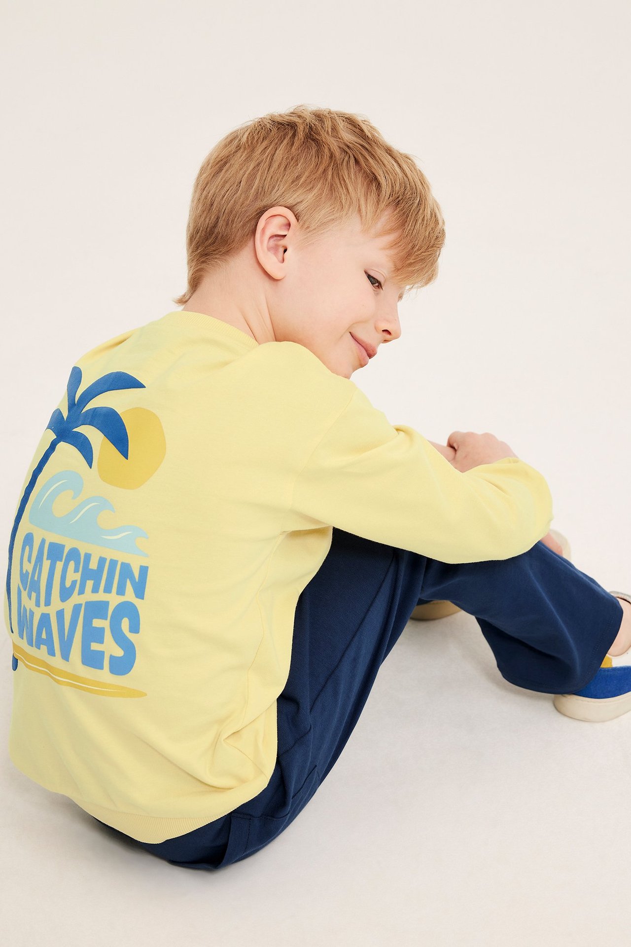 Gult bomullssweatshirt for barn med blått 'Catchin' Waves' ryggtrykk og mørkeblå bukser.