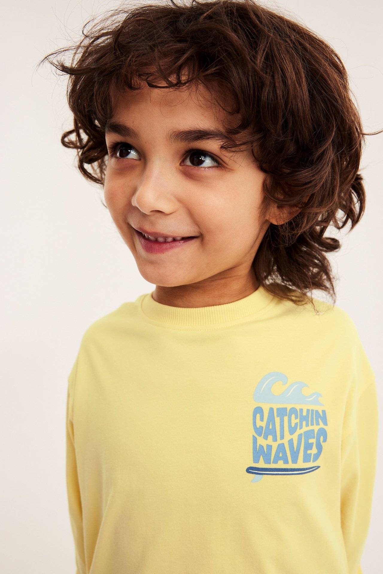 Nærbilde, forfra, av et barn i en gul bomullssweatshirt med blått 'CATCHIN WAVES'-trykk.
