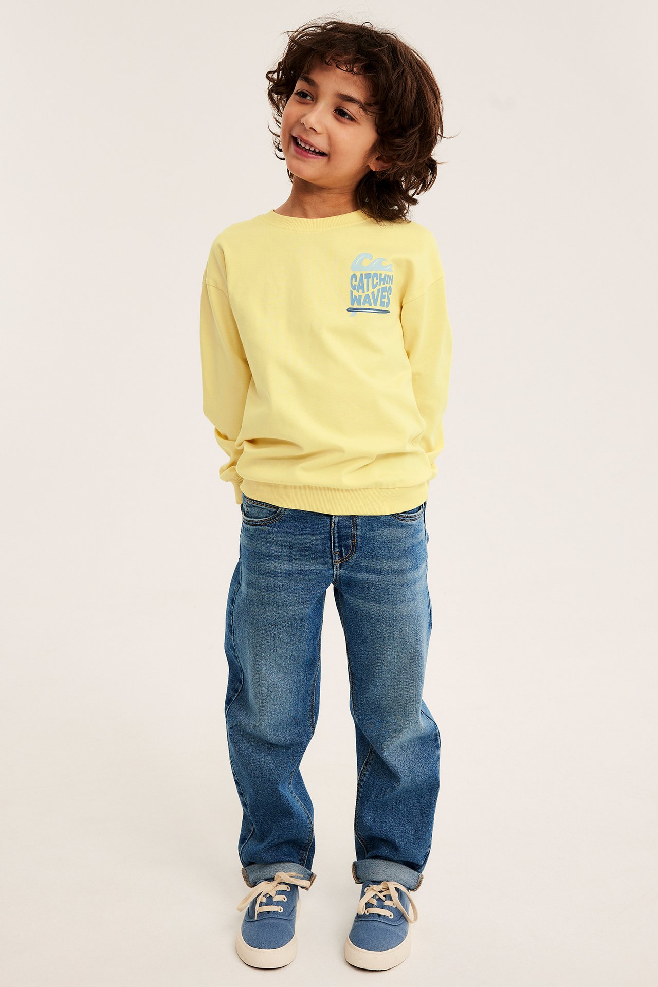 Full frontvisning av et barn i gul bomullssweatshirt med trykk, blå jeans og blå sneakers.