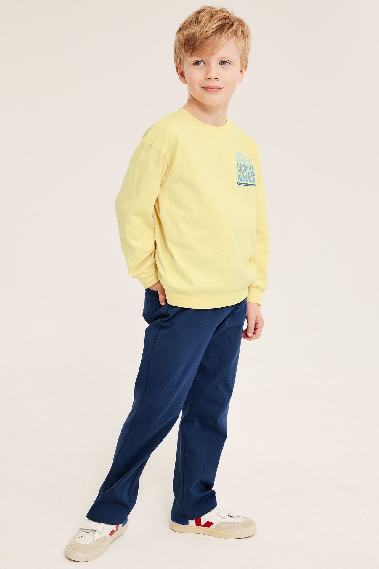 Front-sidevisning av et barn i gul bomullssweatshirt med blått trykk, mørkeblå bukse og hvite joggesko.
