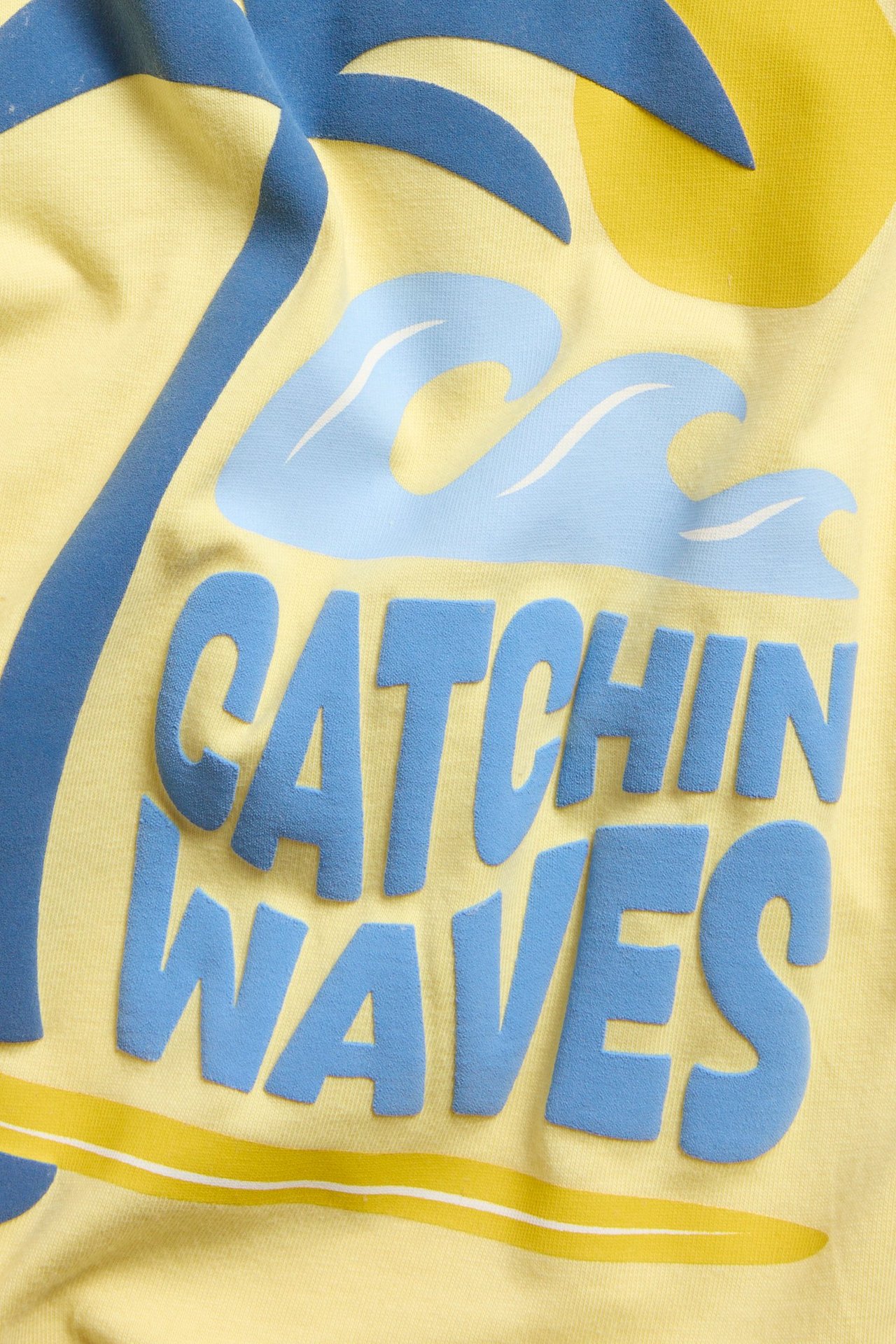 Zbliżenie na żółtą bawełnianą bluzę niemowlęcą z nadrukiem 'Catchin Waves', palmą, falami.
