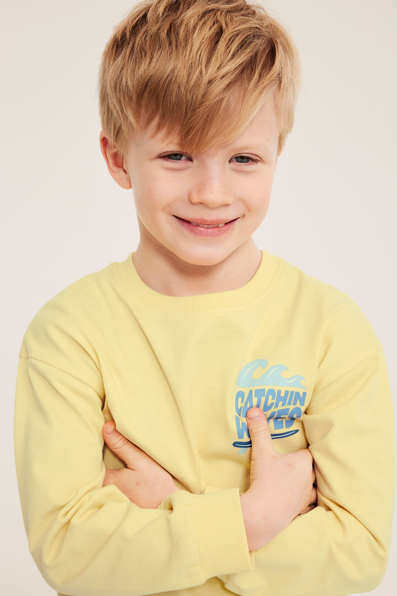 Forfra, smilende barn i en gul bomullssweatshirt med et blått "CATCHIN WAVES" trykk.