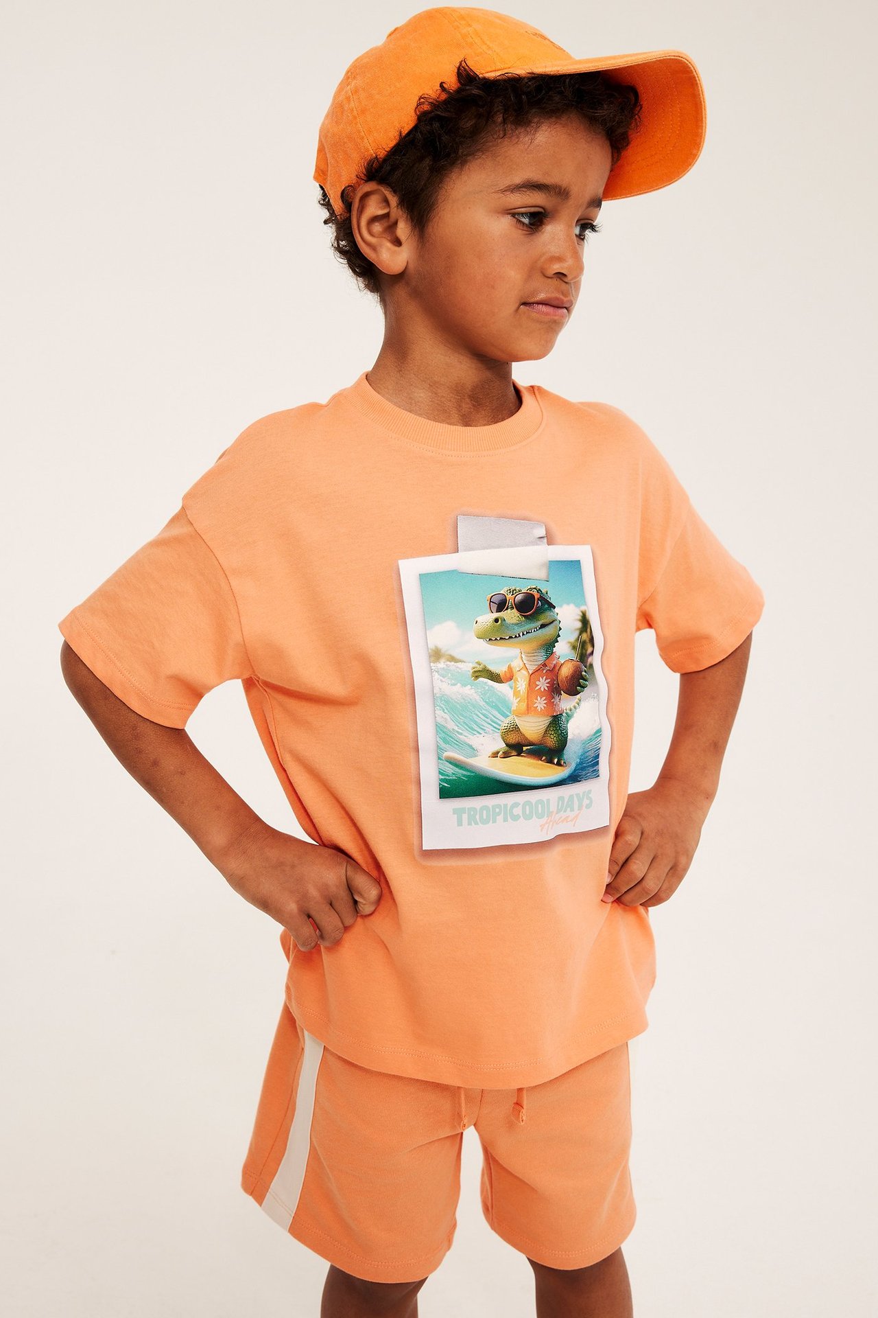 Frontvy av ett barn i en orange bomulls-T-shirt med surfande krokodilprint, orange shorts och keps.