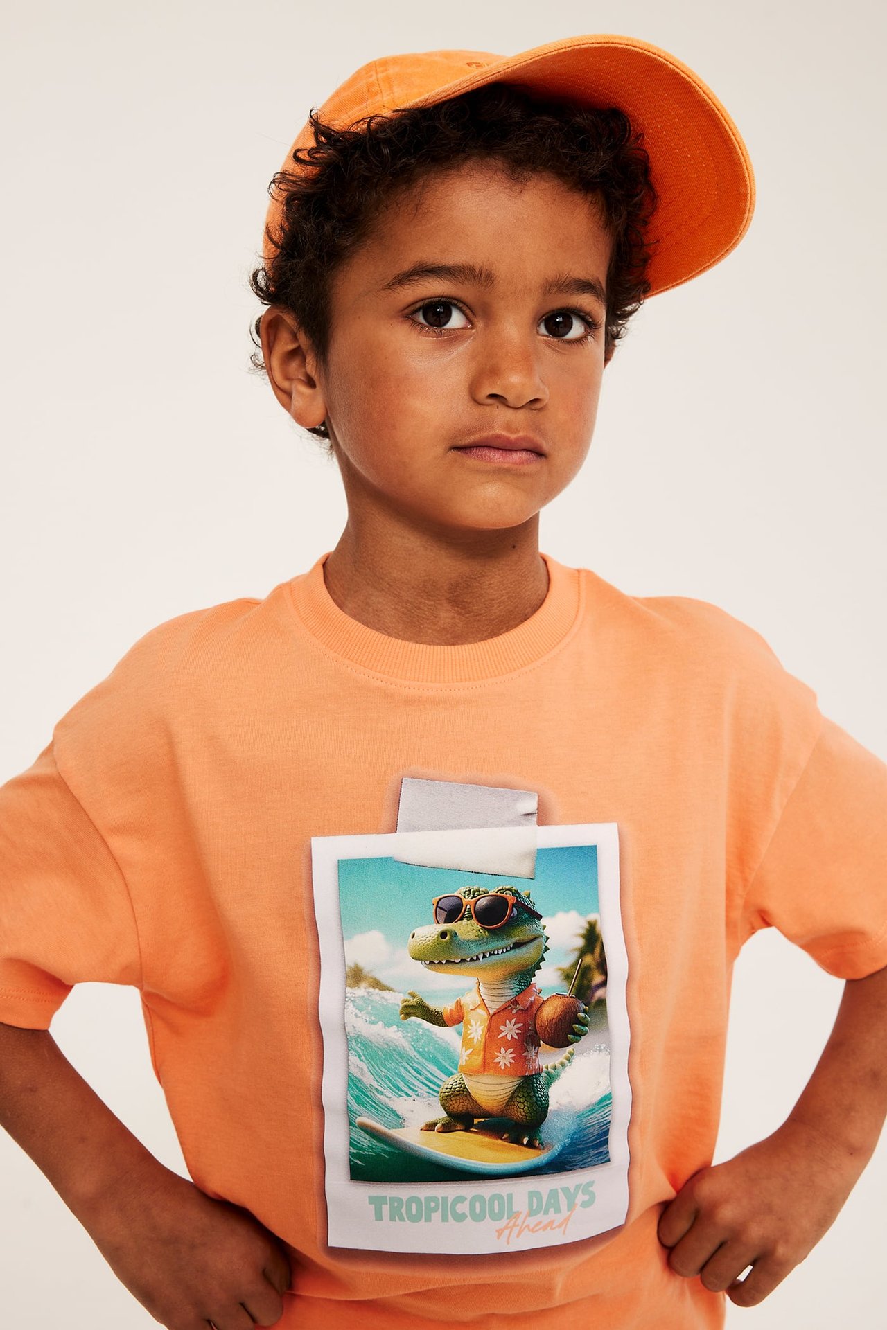 Frontvy av ett barn i en orange bomulls-T-shirt med surfande krokodilprint och en orange keps.