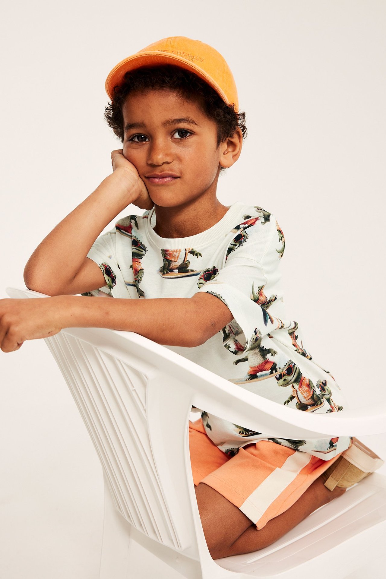 Frontvy: Ett barn bär en off-white t-shirt i bomull med surfande krokodilmönster, orangea shorts och en orange keps.