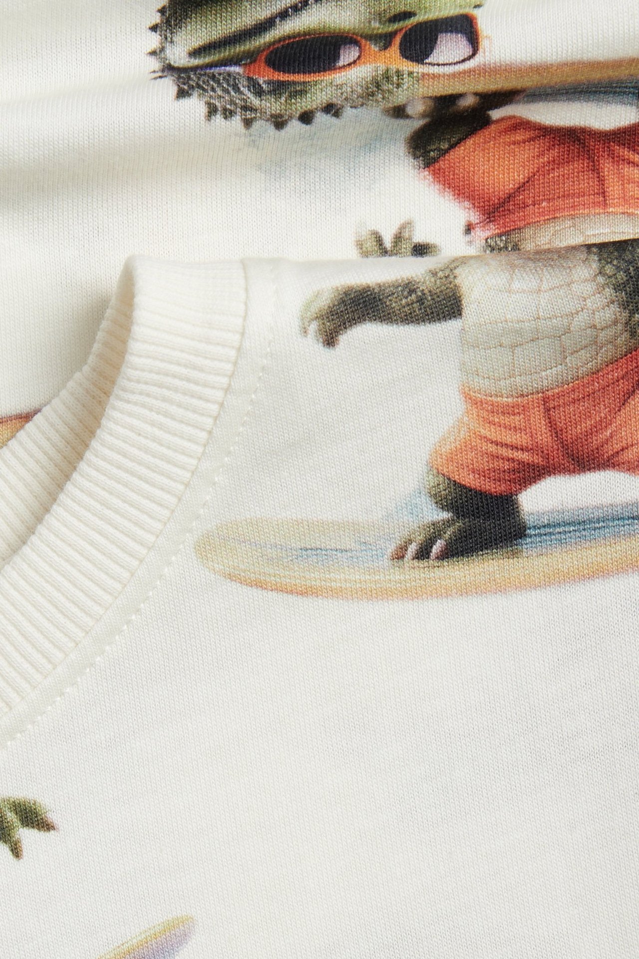 Zbliżenie na kremowobiały bawełniany T-shirt z nadrukiem surfujących krokodyli na całej powierzchni.