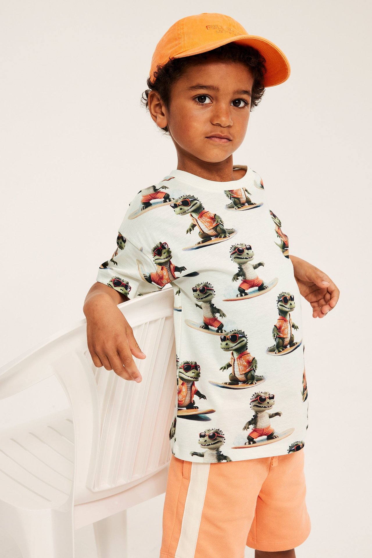 Framifrån: Kid i offwhite bomulls-T-shirt med mönster av surfande krokodiler, orange shorts och orange keps.