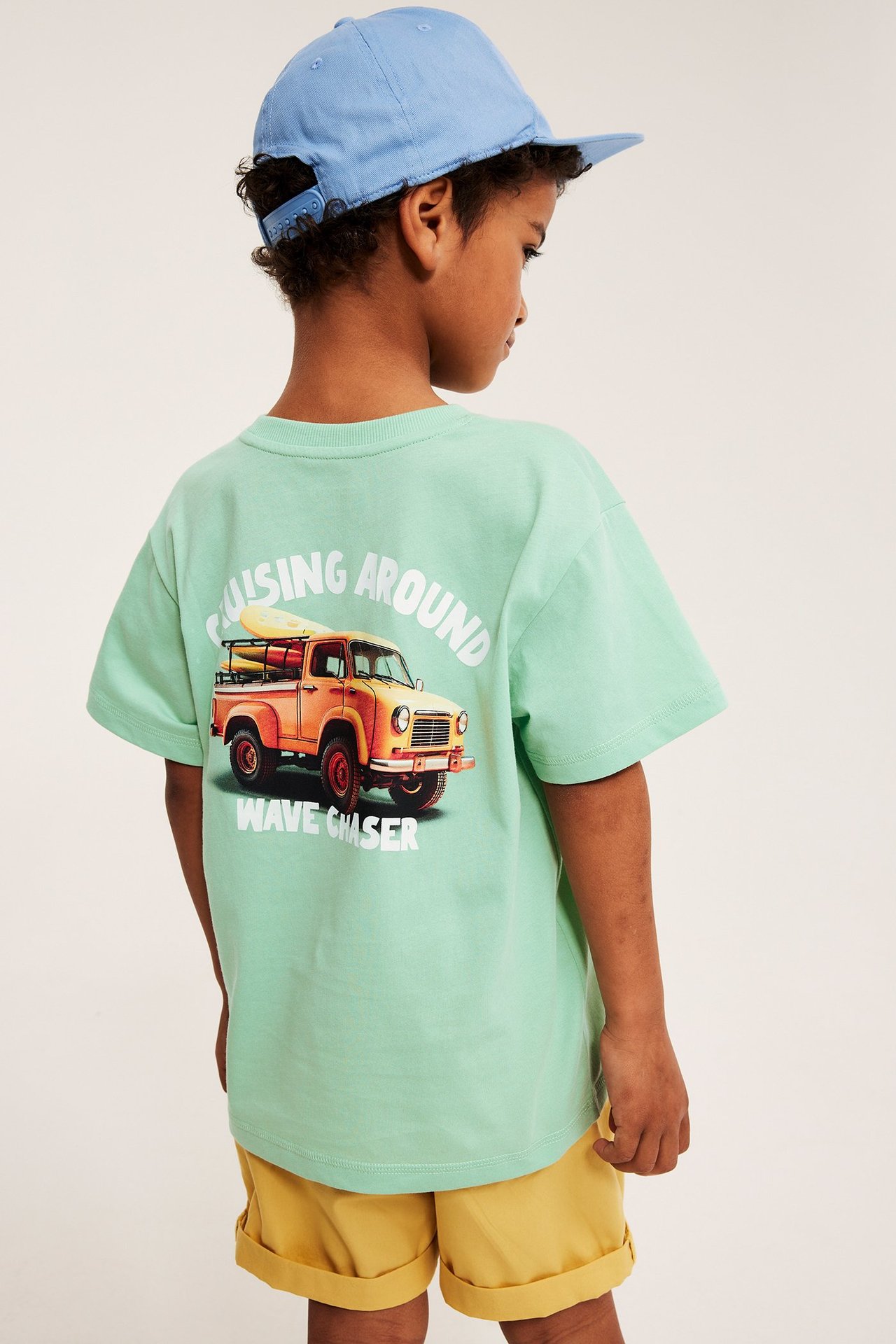 Lapsi selin, ylisuuri vaaleanvihreä t-paita autokuviolla, keltaiset shortsit, sininen lippis.