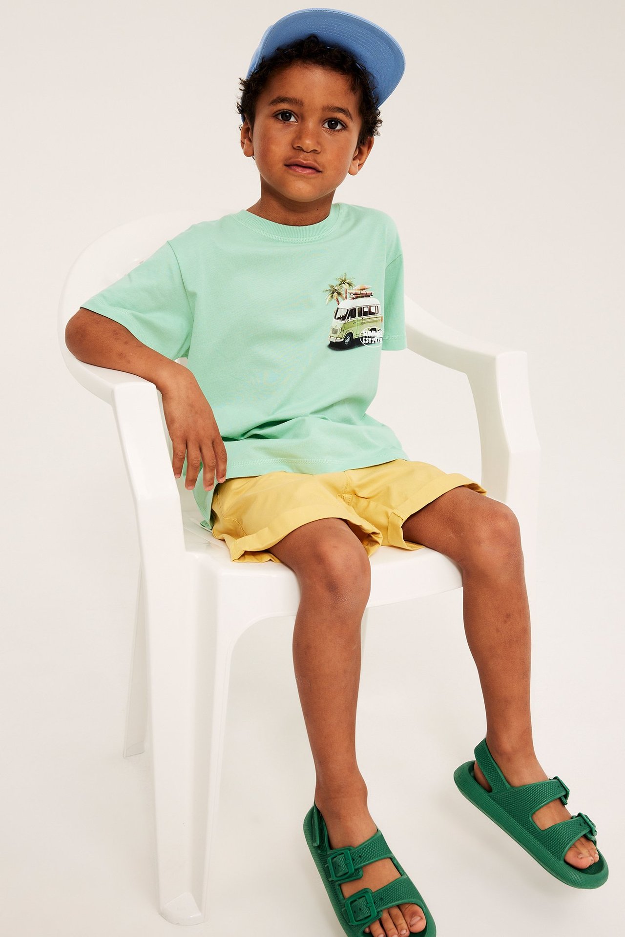 Pojke i grön oversize t-shirt med van-tryck, gula shorts, gröna sandaler och blå keps.