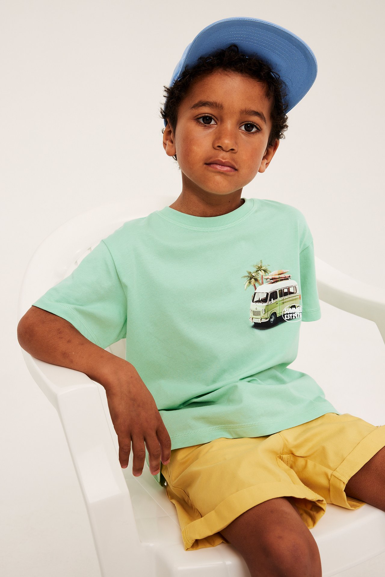 Lapsi edestäpäin vihreässä oversized T-paidassa autokuvalla, keltaisissa shortseissa ja sinisessä lippalakissa.