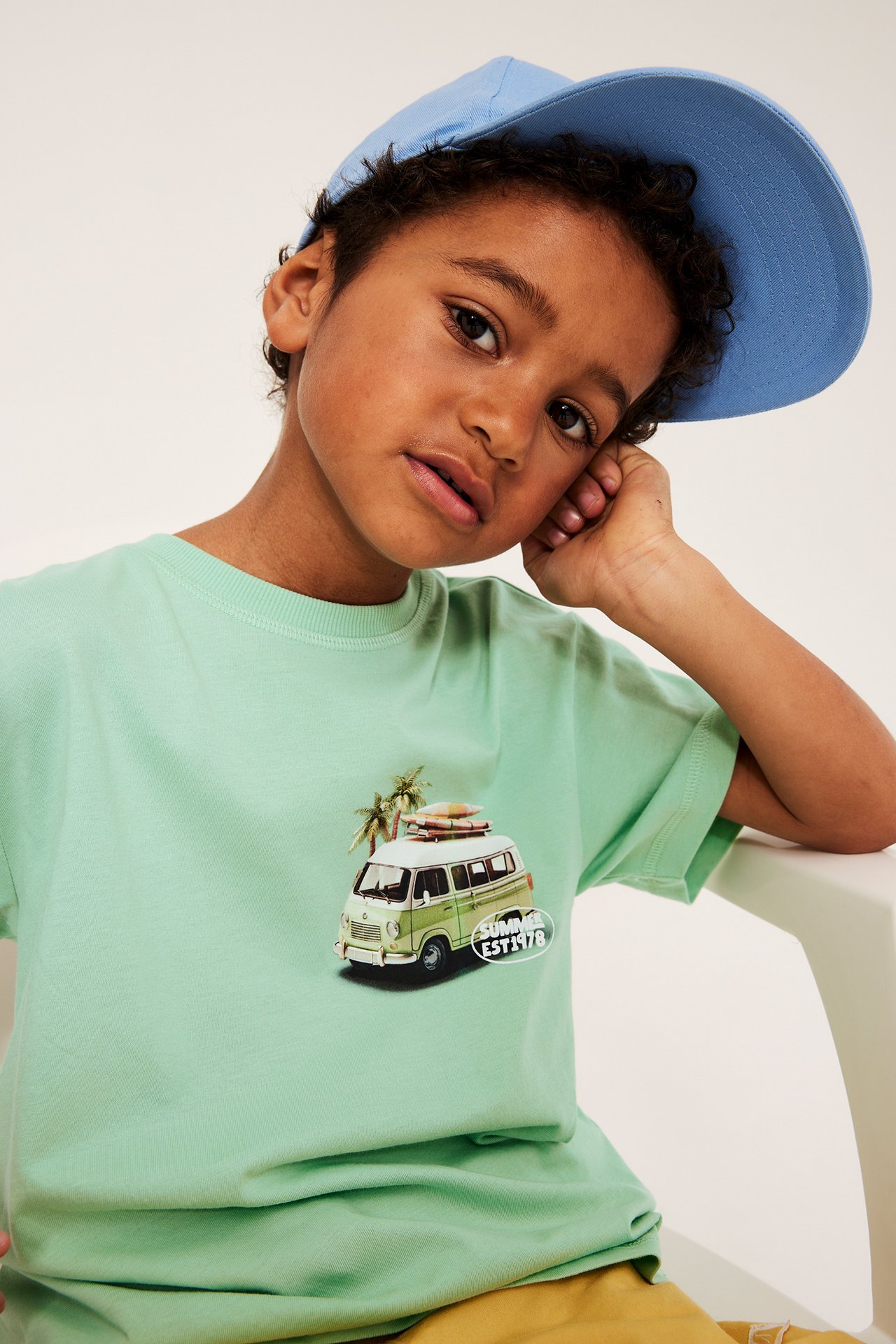 Närbild framifrån på ett barn i en ljusgrön oversize bomulls-t-shirt med bilmotiv och en blå keps.