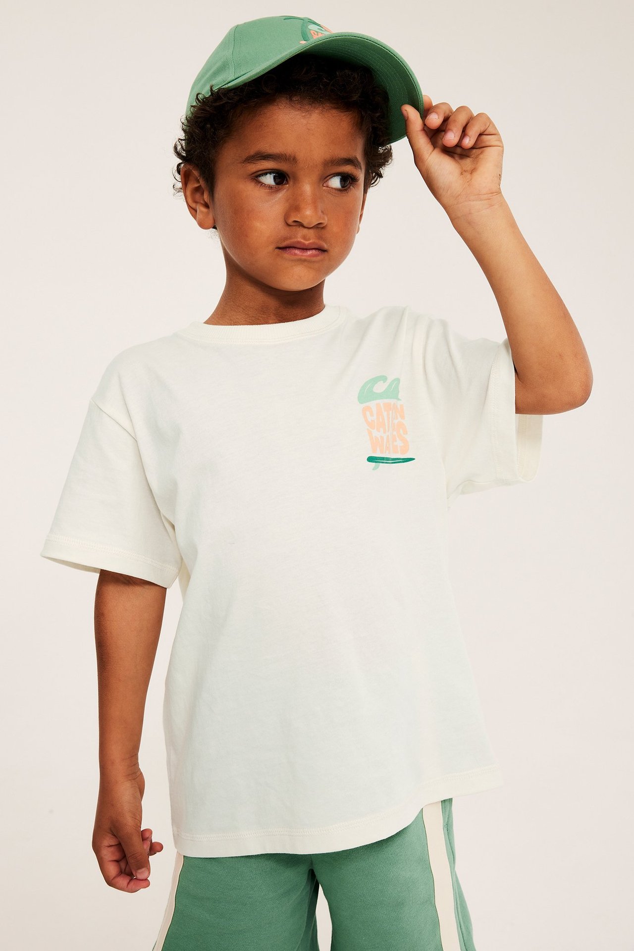Frontvy: Barn i offwhite oversize bomullst-shirt med "Catch Waves"-tryck, gröna shorts och grön keps.
