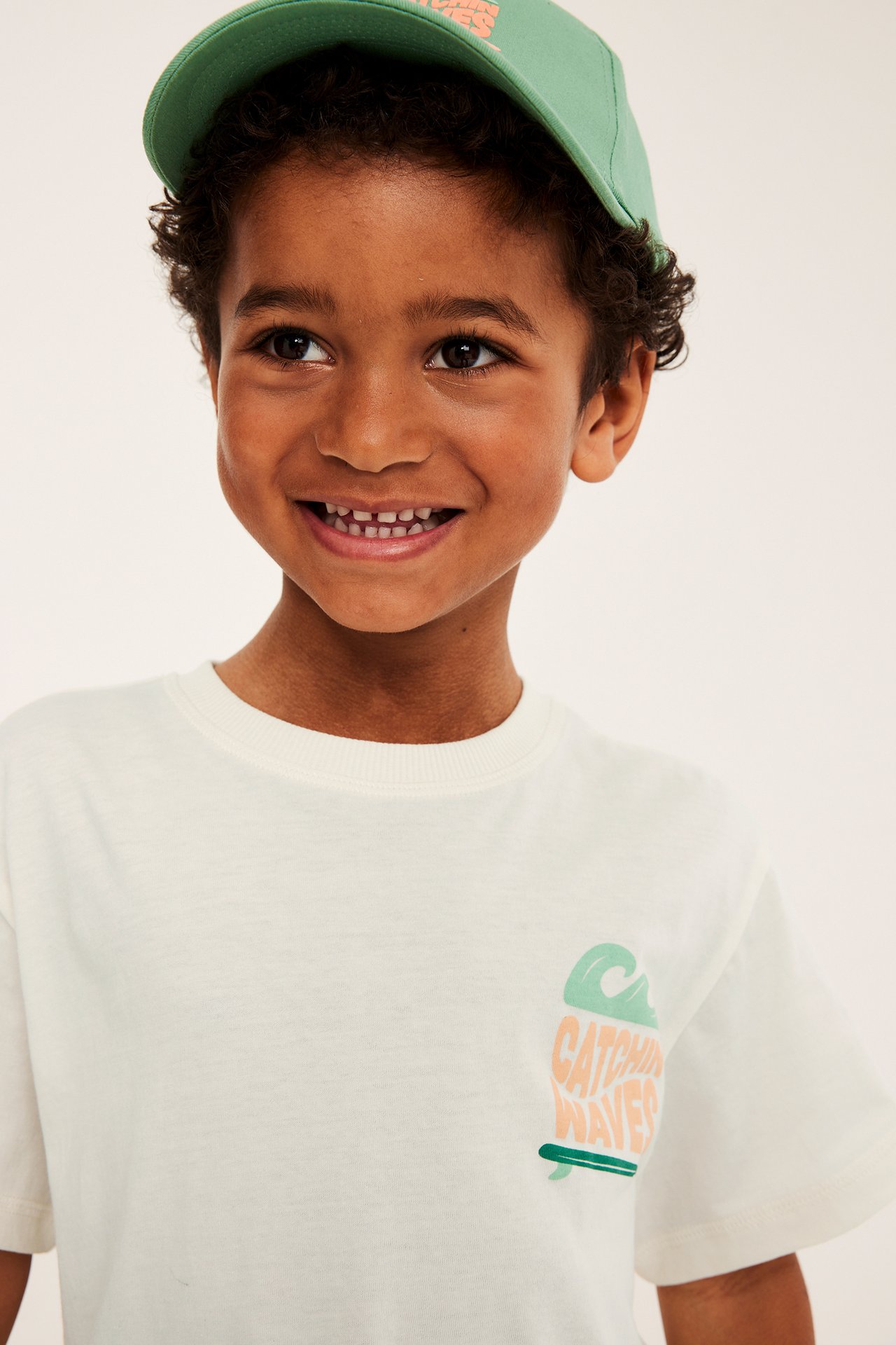 Hymyilevä lapsi, edestä, luonnonvalkoisessa oversized puuvilla t-paidassa 'Catchin' Waves' -printillä ja vihreässä lakissa.
