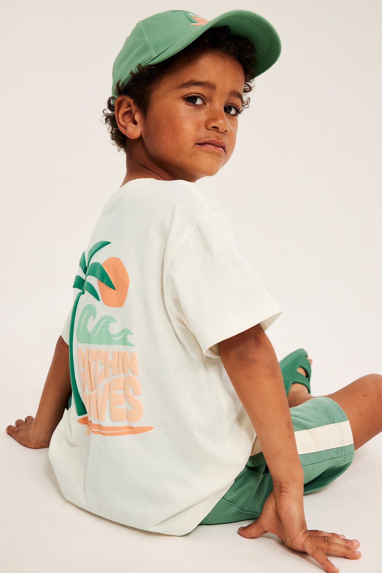 Dziecko od tyłu: T-shirt oversize off-white z plażowym nadrukiem, zielone szorty i czapka.