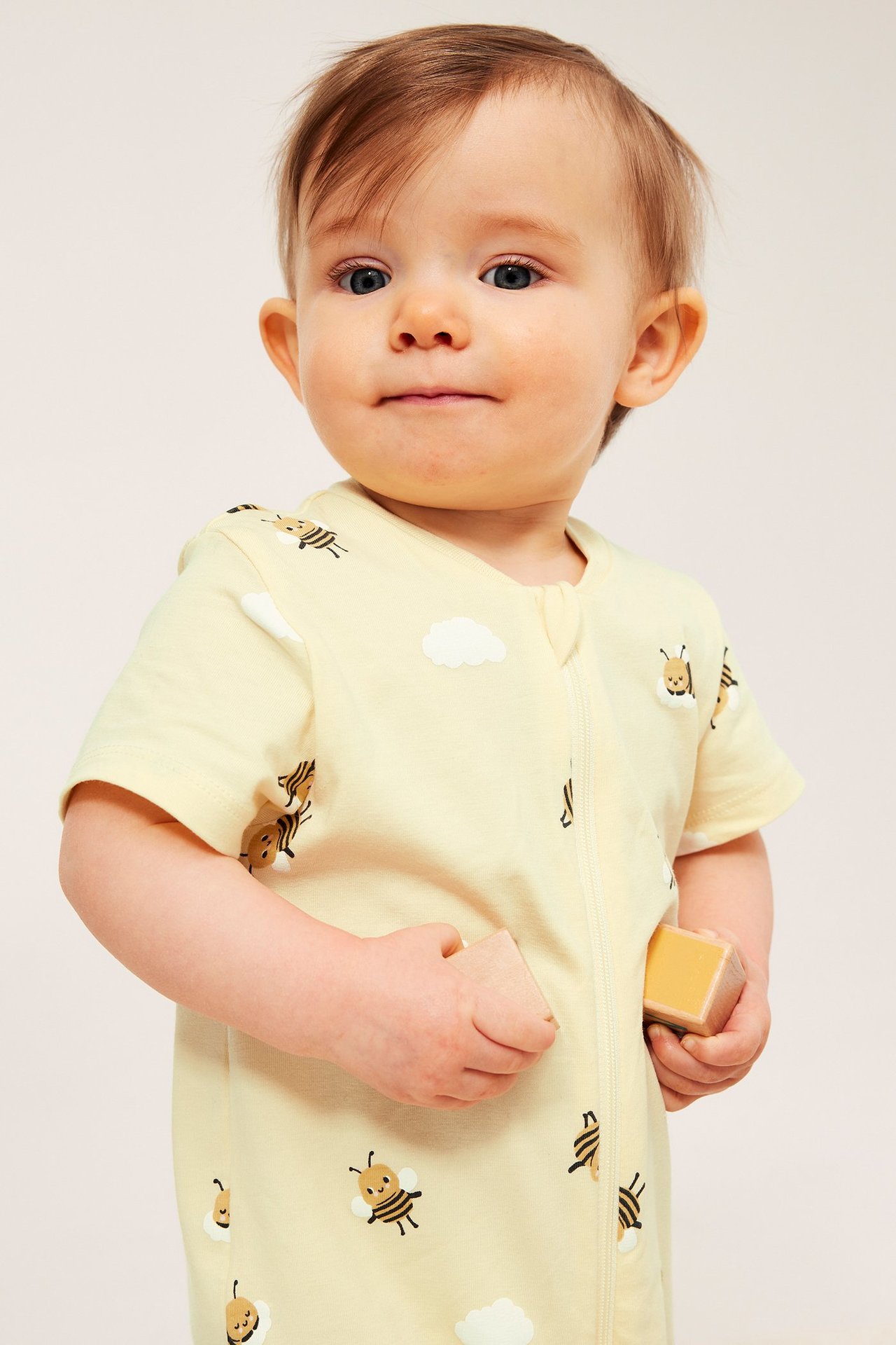 Framifrån, en baby i ljusgul kortärmad romper med bi-mönster, håller träklossar.