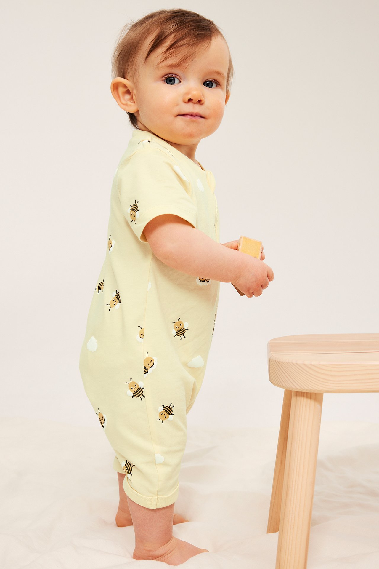 Sivukuva: Vauva vaaleankeltaisessa lyhythihaisessa haalarissa mehiläiskuviolla, pitäen puupalikkaa.