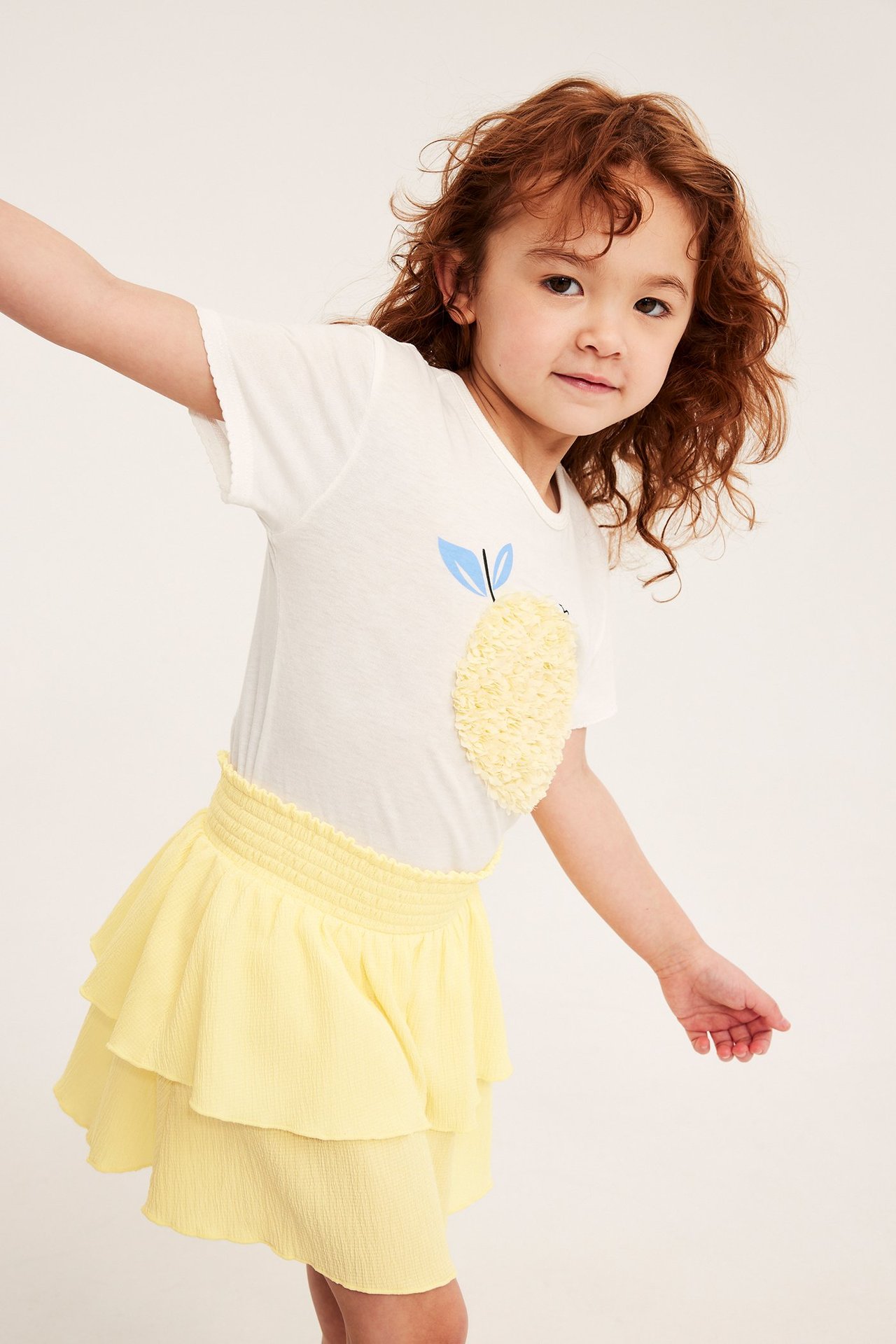 Widok z przodu: dziecko w jasnej, żółtej, marszczonej spódniczce z falbanami i białej koszulce z cytryną.