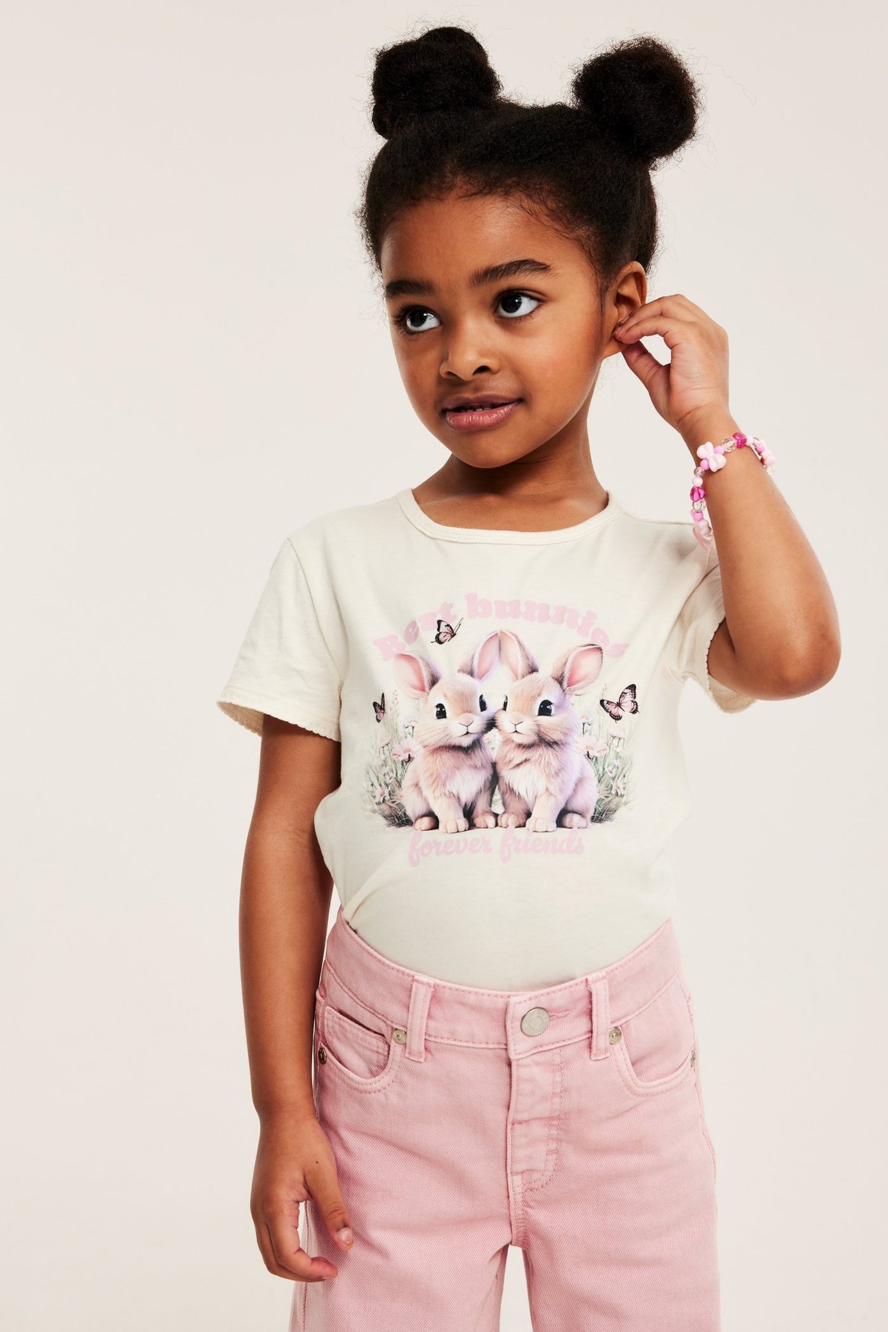 Edestäpäin lapsi yllään luonnonvalkoinen puuvilla t-paita kanipainatuksella, pinkit farkut ja vaaleanpunainen rannekoru.