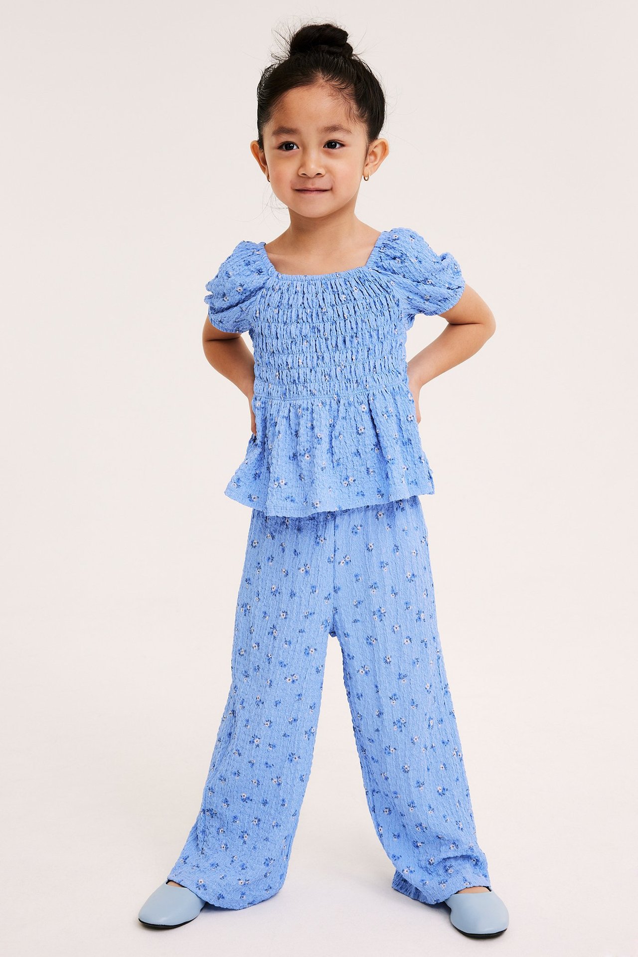 Framifrån, ett barn i ljusblå blommiga strukturerade culottebyxor och en matchande smocktopp.