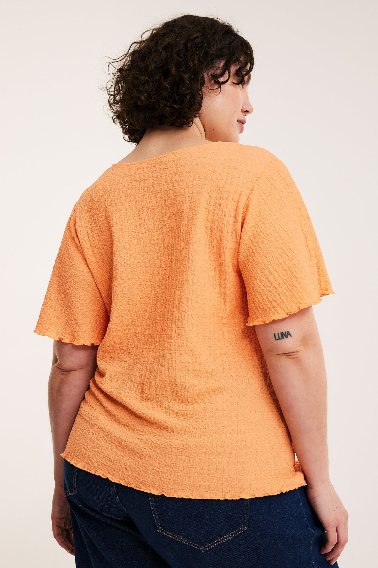Ryggvy av kvinna i en lös, orange kortärmad topp med struktur och mörkblå jeans.