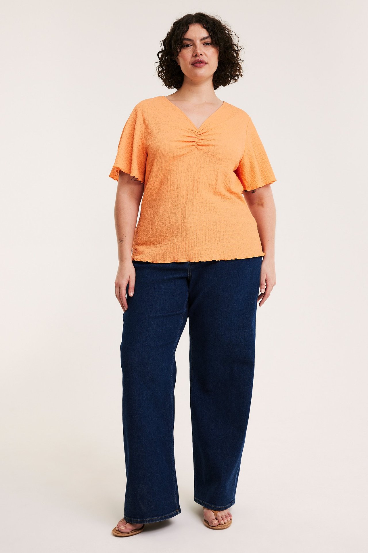 Framifrån, en kvinna bär en orange texturerad V-ringad topp, mörkblå vida jeans och platta sandaler.