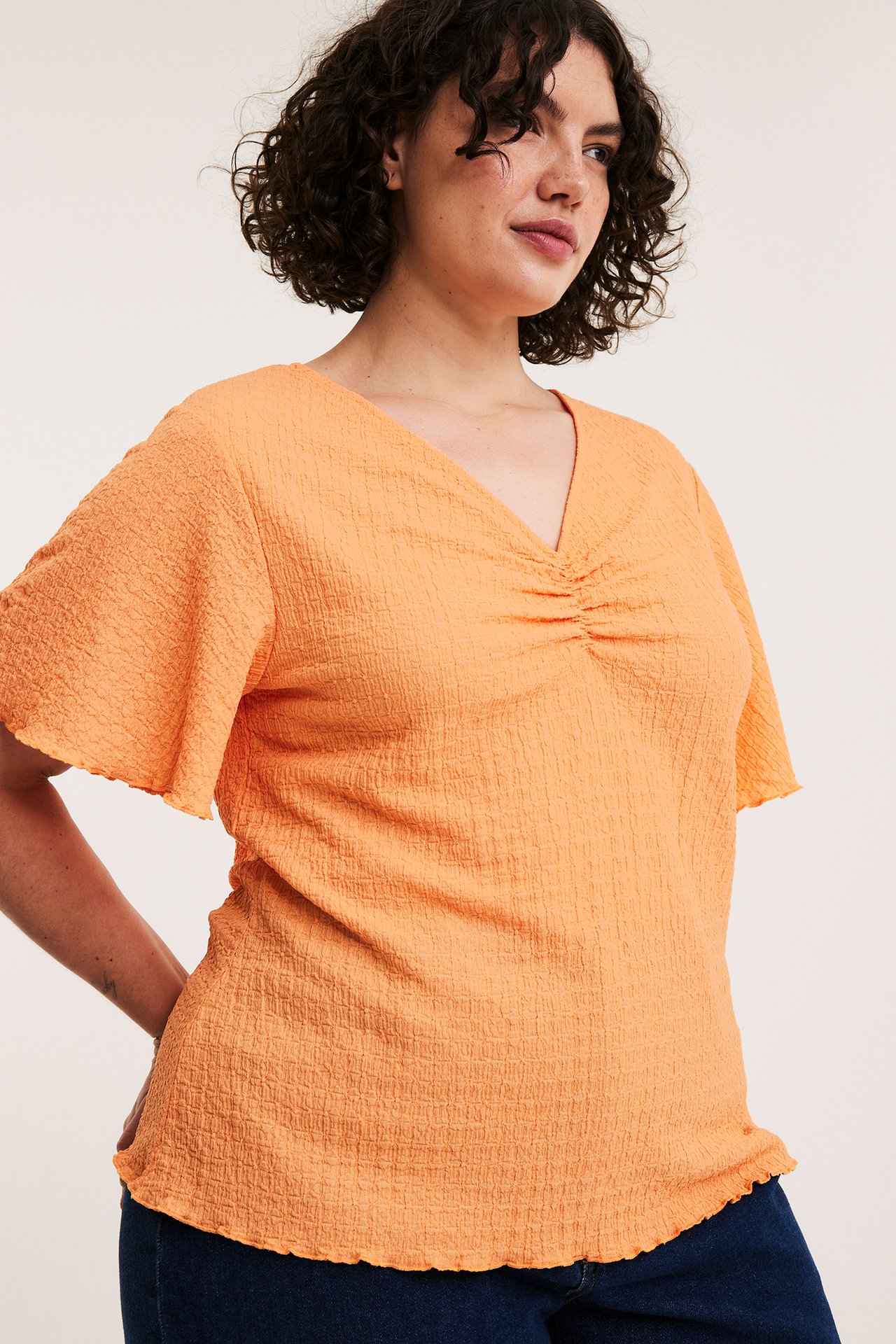 Framifrån, kvinna i orange strukturerad V-ringad topp med rynkad detalj och mörkblå jeans.