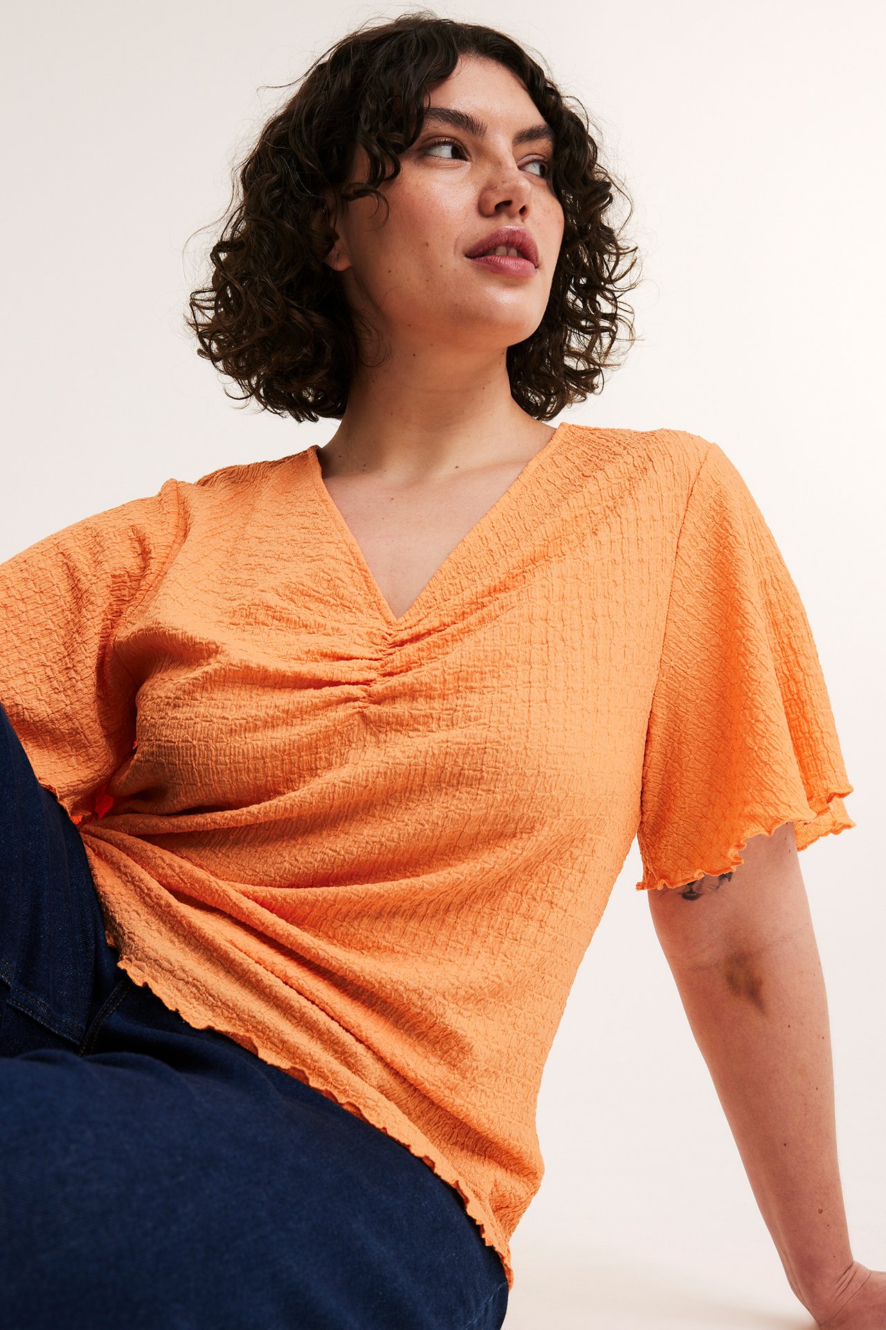 Frontvy av kvinna i orange strukturerad V-ringad topp med volangärmar och mörkblå jeans.