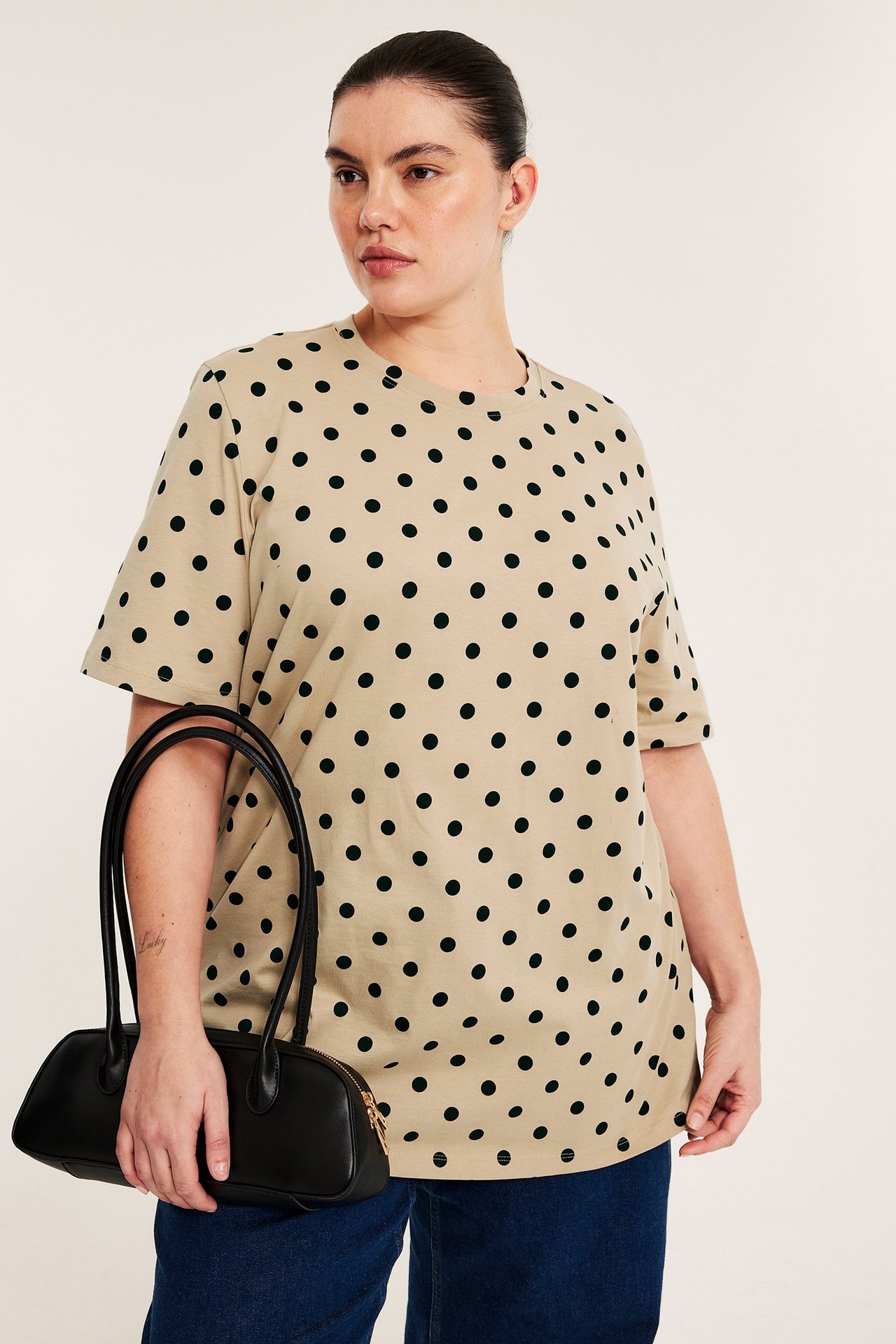 Forfra: kvinne i en beige prikkete lang t-skjorte i bomull, mørkeblå bukser, holder en svart håndveske.