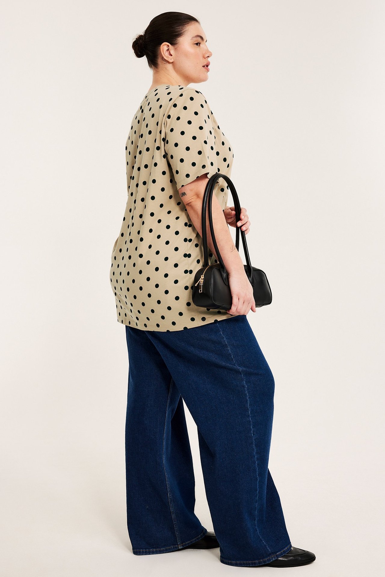 Sidebilde: kvinne i beige prikkete lang t-skjorte, mørkeblå vide jeans, holder svart veske.