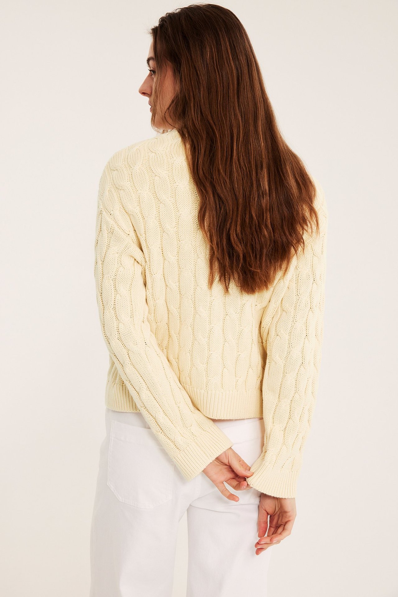 Kobieta tyłem-bokiem, w jasnożółtym swetrze z warkoczami i białych spodniach, ręce z tyłu.