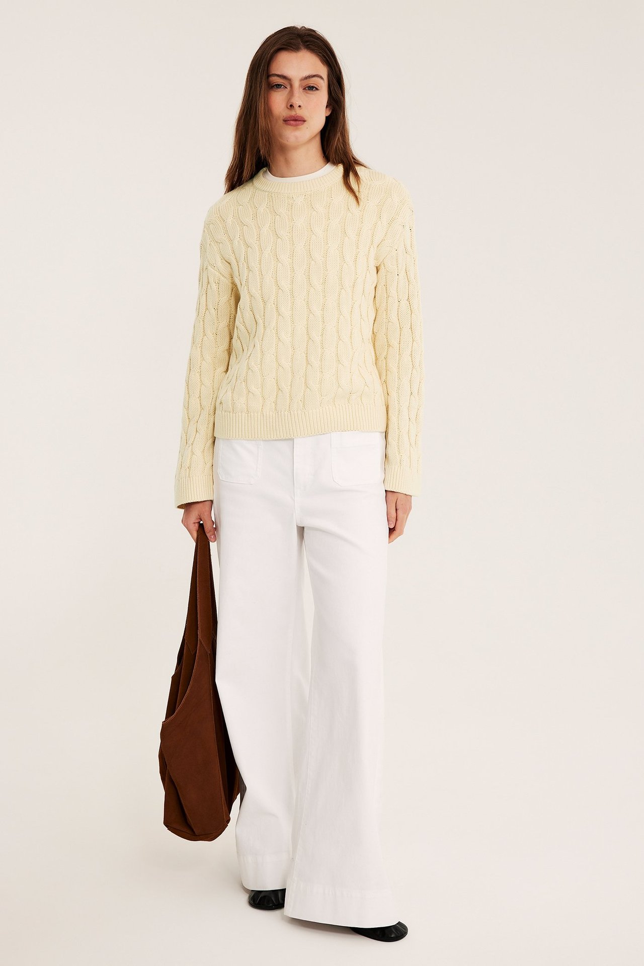 Kvinna i ljusgul kabelstickad tröja, vita vida byxor, svarta skor, håller brun tygväska.