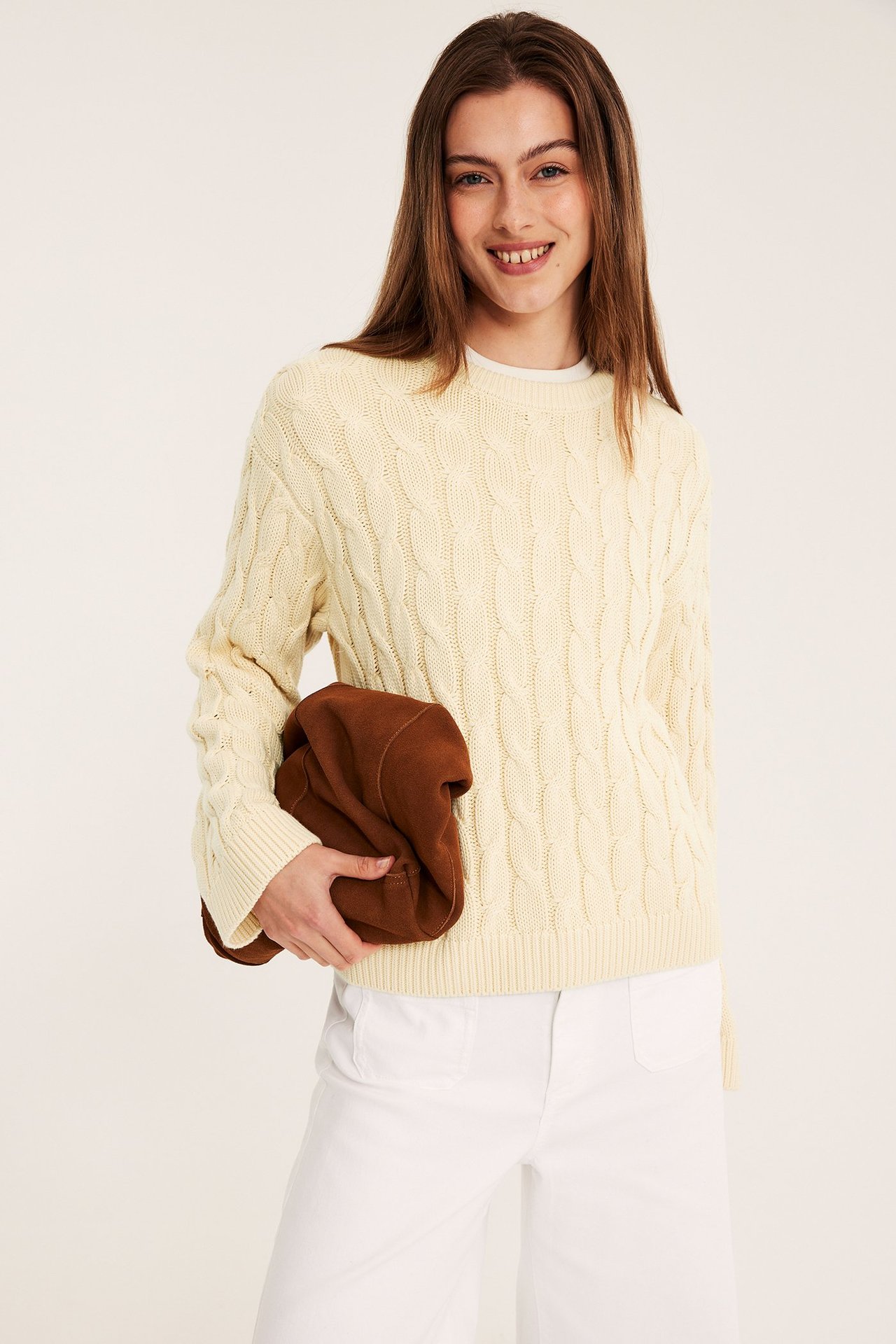 Etunäkymä: nainen vaaleankeltaisessa palmikkoneuleessa, valkoisissa leveissä housuissa, pitää ruskeaa pussukkaa.