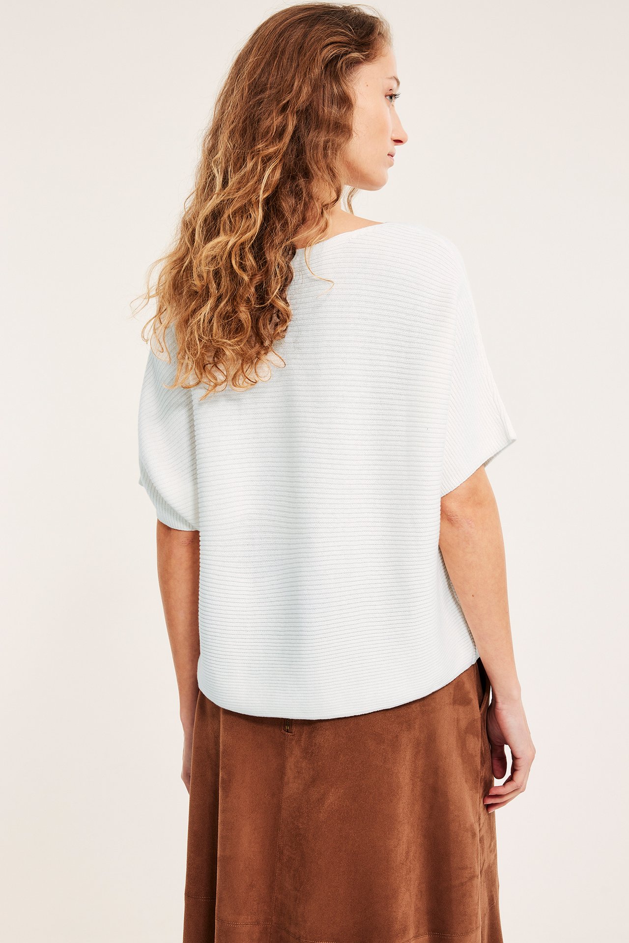 Bakfra: kvinne i en løs hvit ribbestrikket kortermet genser og et brunt A-formet skjørt.