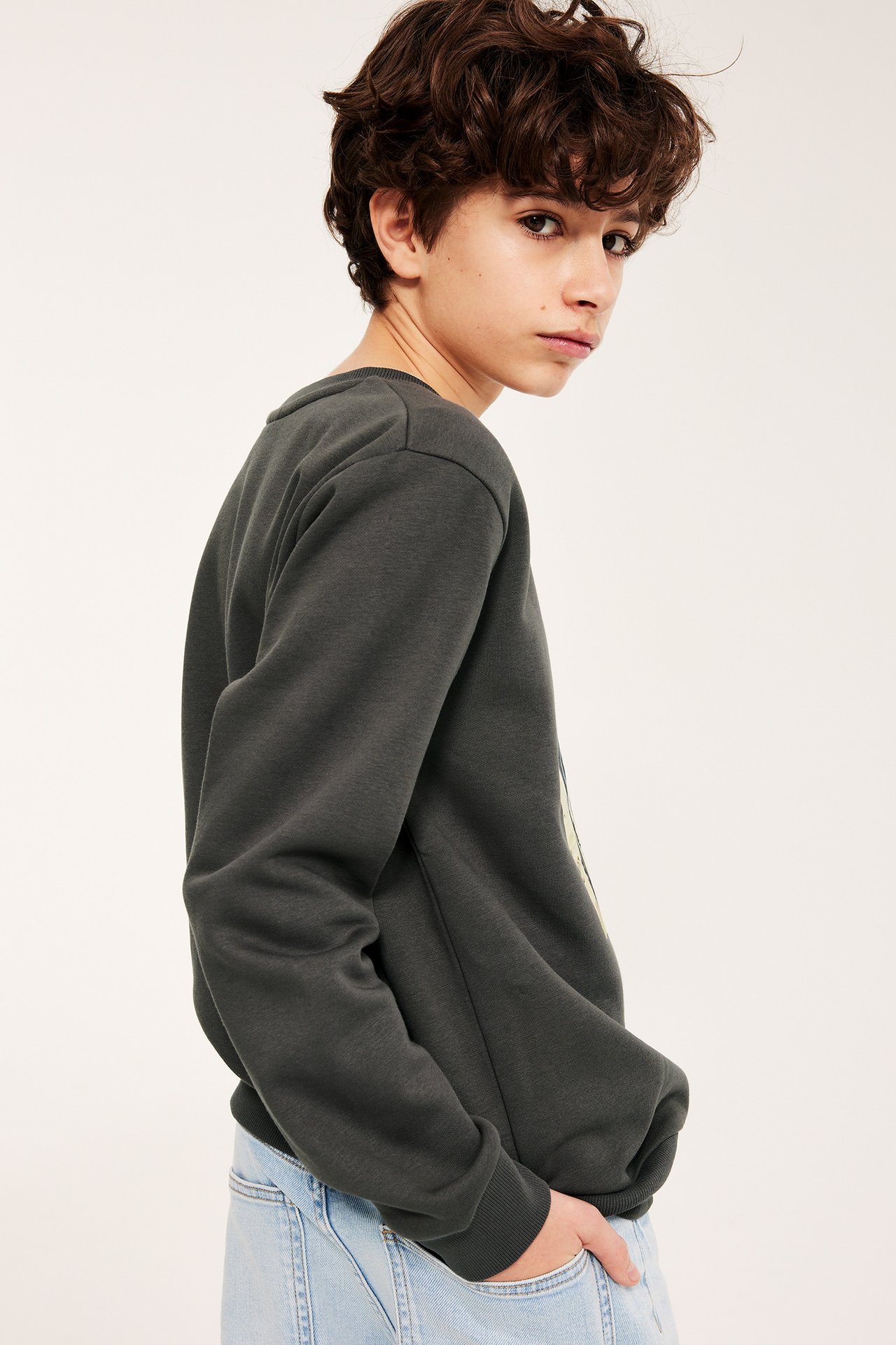 Widok z boku: Dziecko w ciemnoszarej bluzie z nadrukiem i jasnoniebieskich dżinsach.