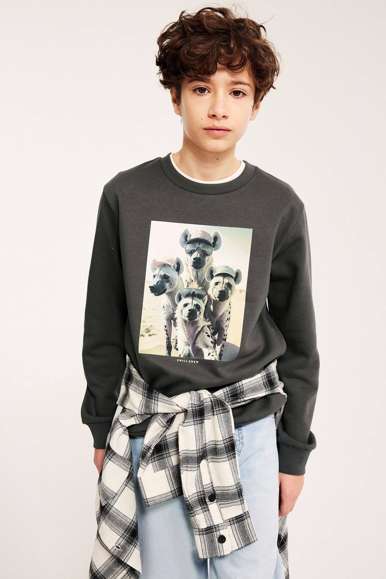Frontutsikt, barn i svart sweatshirt med hyenatrykk, hvit t-skjorte, lyseblå jeans og rutete skjorte knyttet.