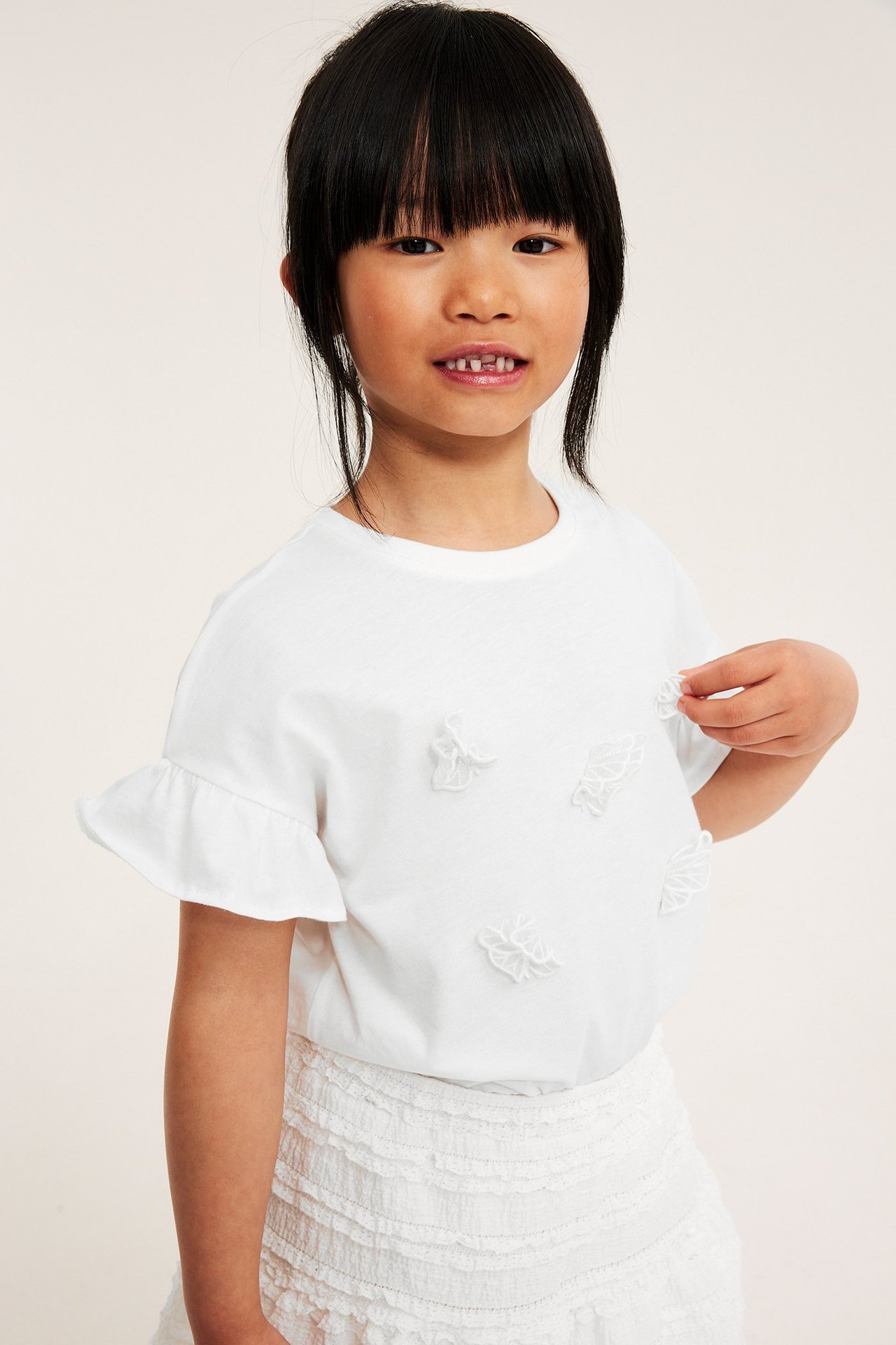 Frontvy: Barn i offwhite bomullst-shirt med volangärmar, 3D-fjärilsapplikationer, rör vid en.