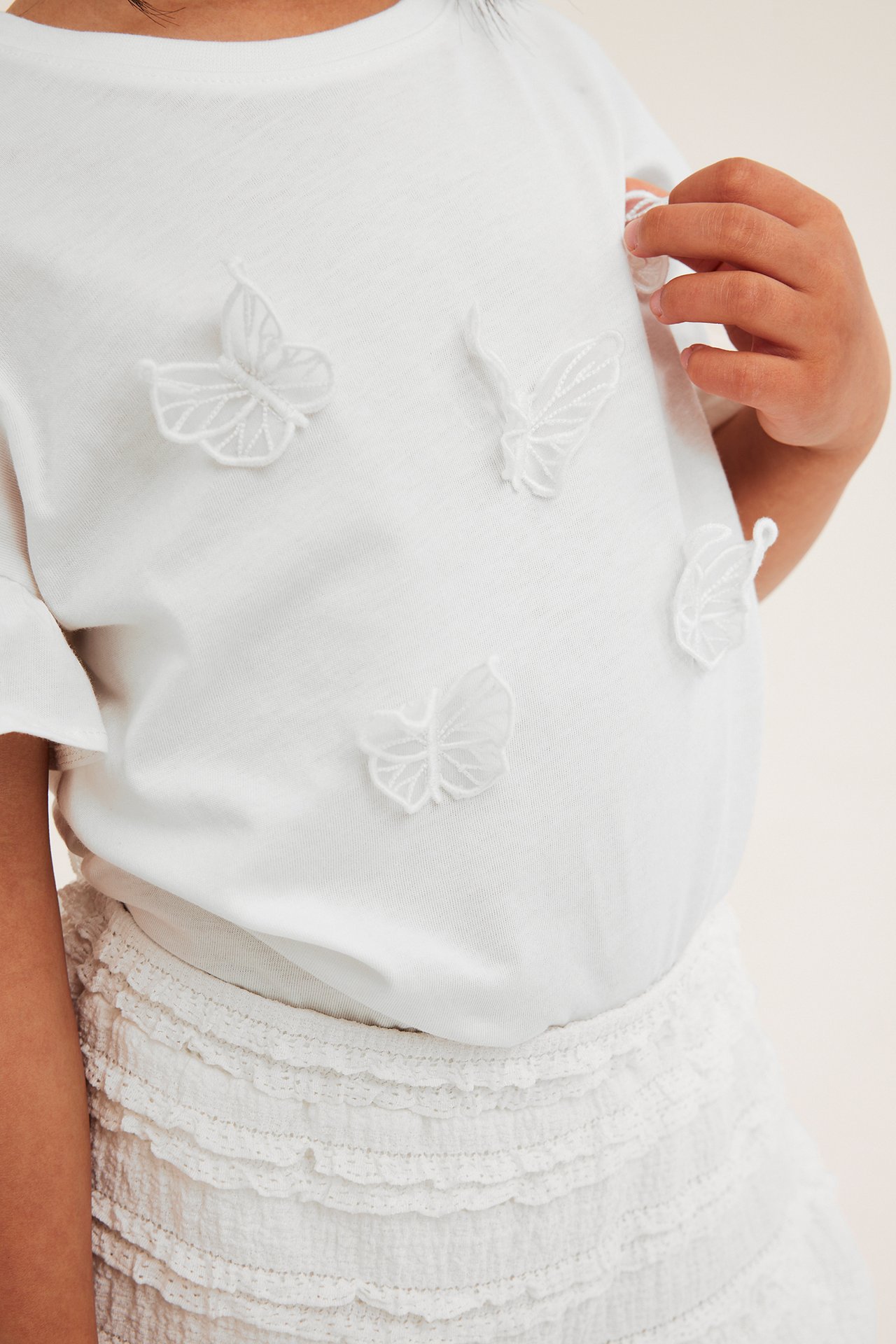 Forfra: Barns off-white bomulls-T-skjorte med 3D sommerfuglapplikasjoner; hånd berører en. Hvit volangskjørt.
