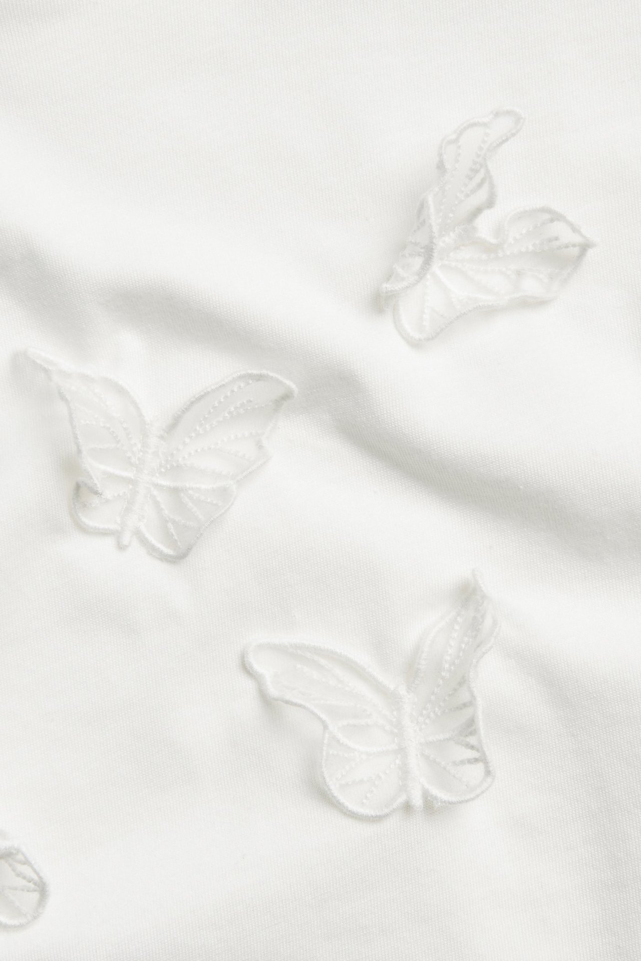 Zbliżenie na ecru bawełniany top z delikatnymi aplikacjami 3D w kształcie motyli z siateczki.