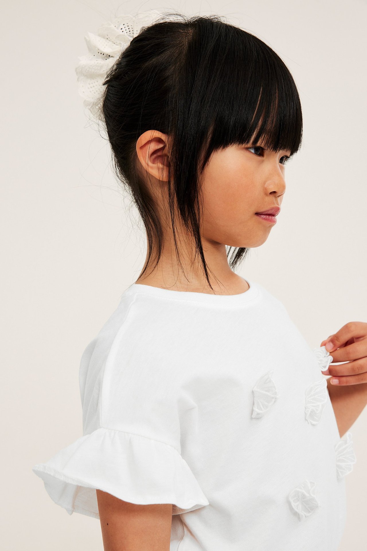 Sivukuva: Lapsi luonnonvalkoisessa t-paidassa röyhelöhihoilla, mesh-perhosilla ja valkoisella hiusasusteella.