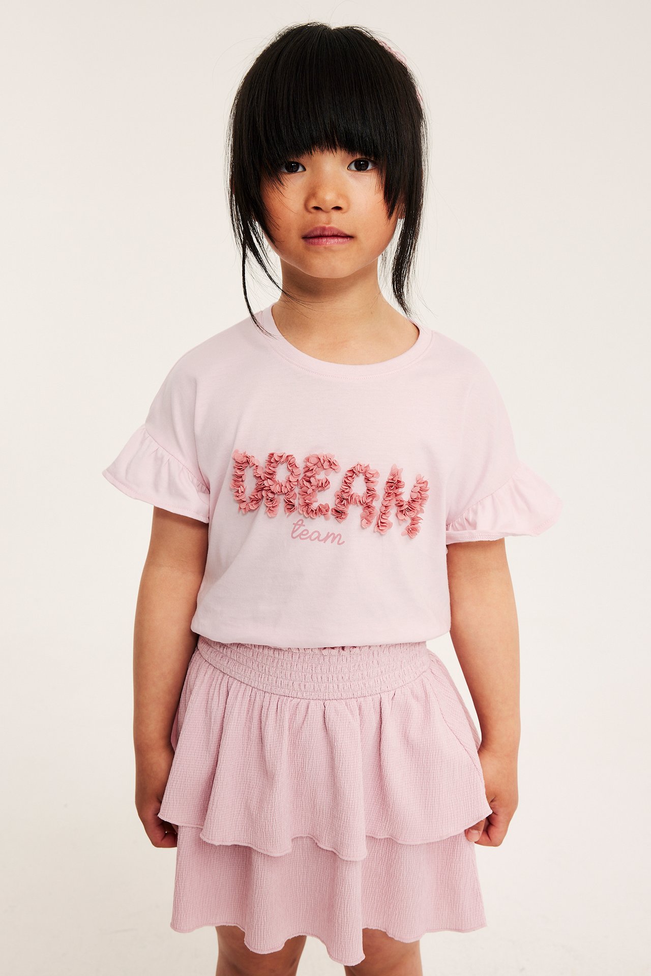 Lapsi yllään vaaleanpunainen T-paita röyhelöhihoilla, 'DREAM team' -verkkokuvio ja kerroksellinen hame. Edestäpäin.
