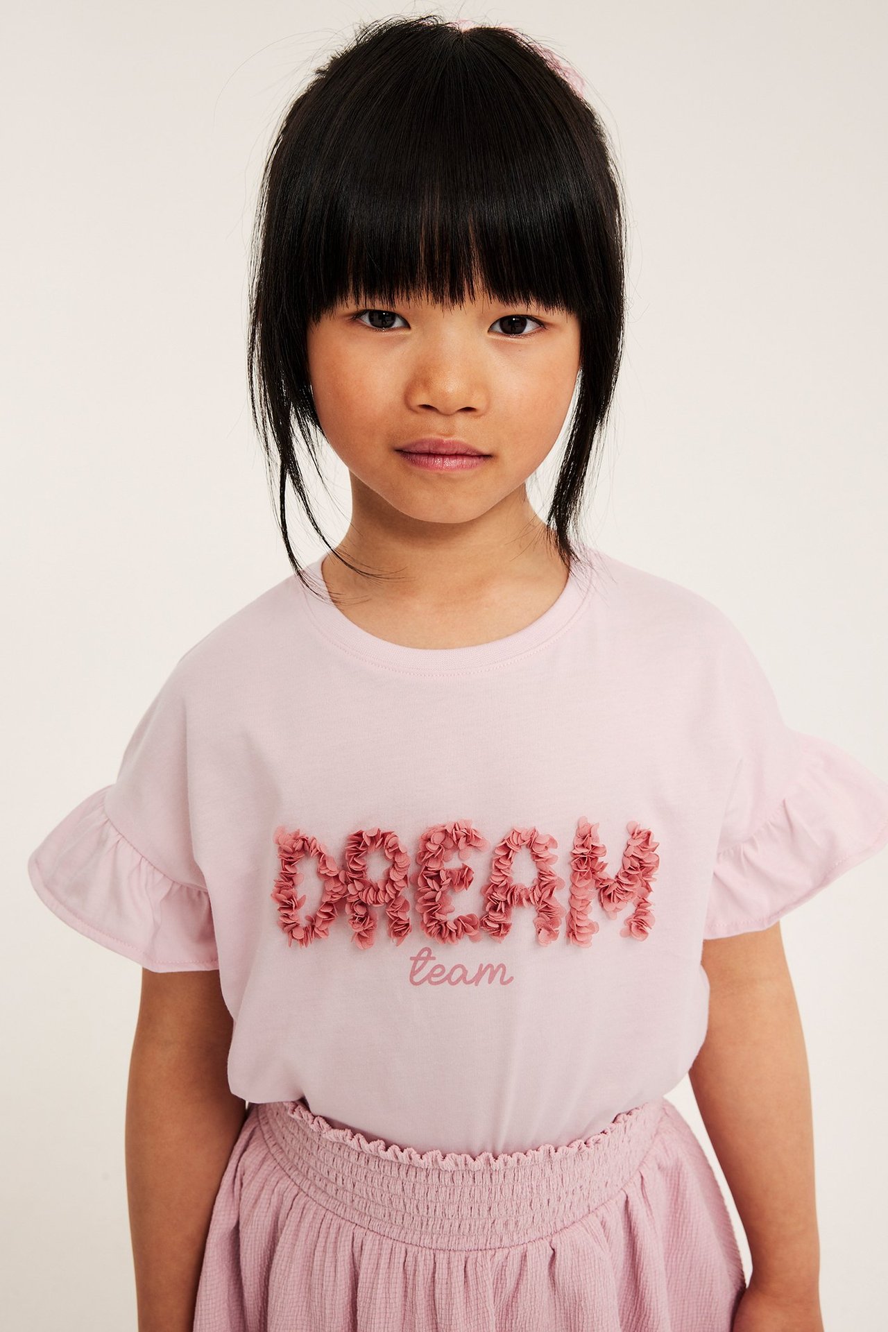Jasnoróżowa koszulka z falbankami na rękawach i różową aplikacją 'DREAM team' z siateczki, noszona przez dziecko z różową spódnicą.