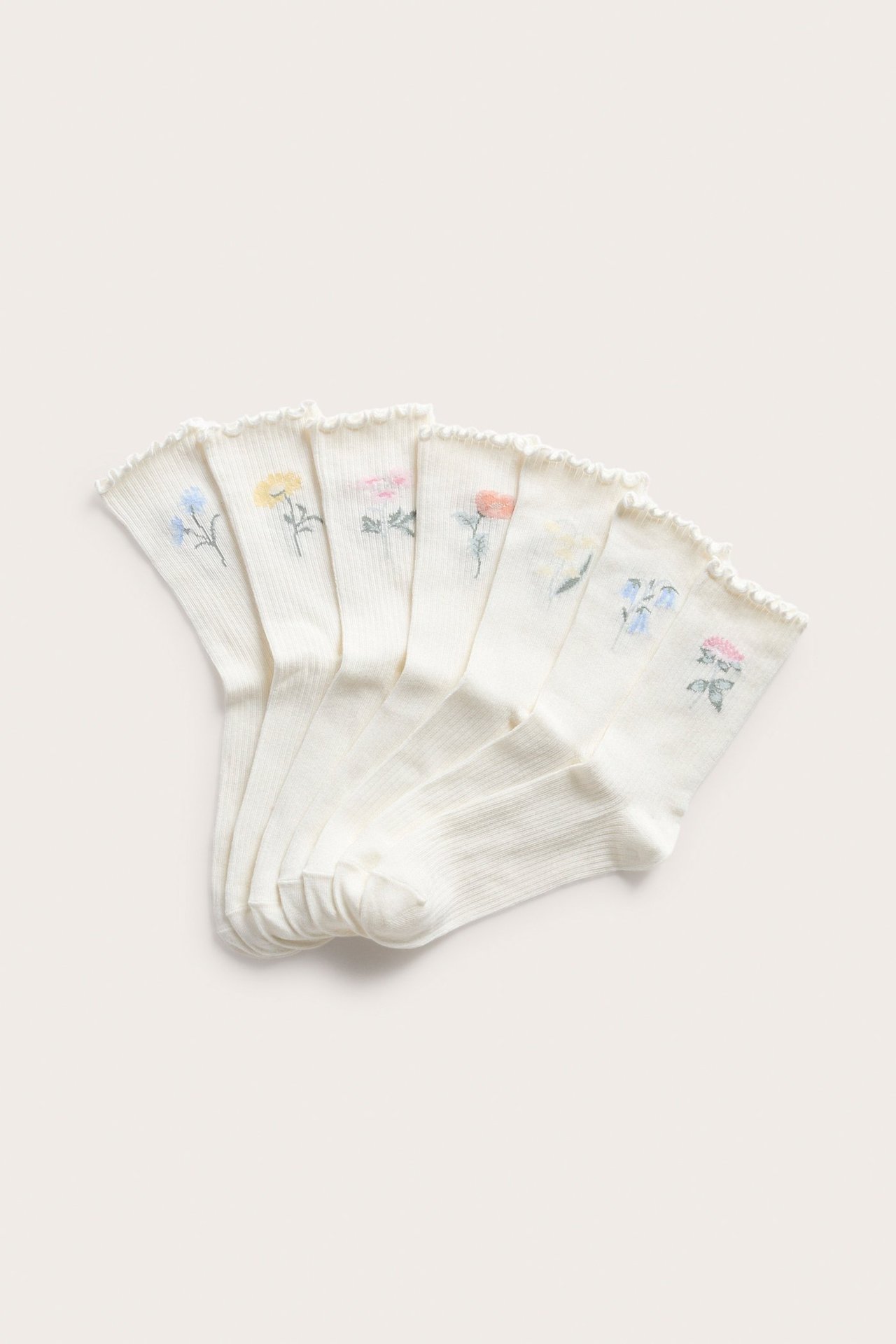 Ylhäältä näkymä seitsemästä luonnonvalkoisesta ribatusta sukasta, joissa on röyhelöreunat ja värikkäitä kukkakirjailuja.