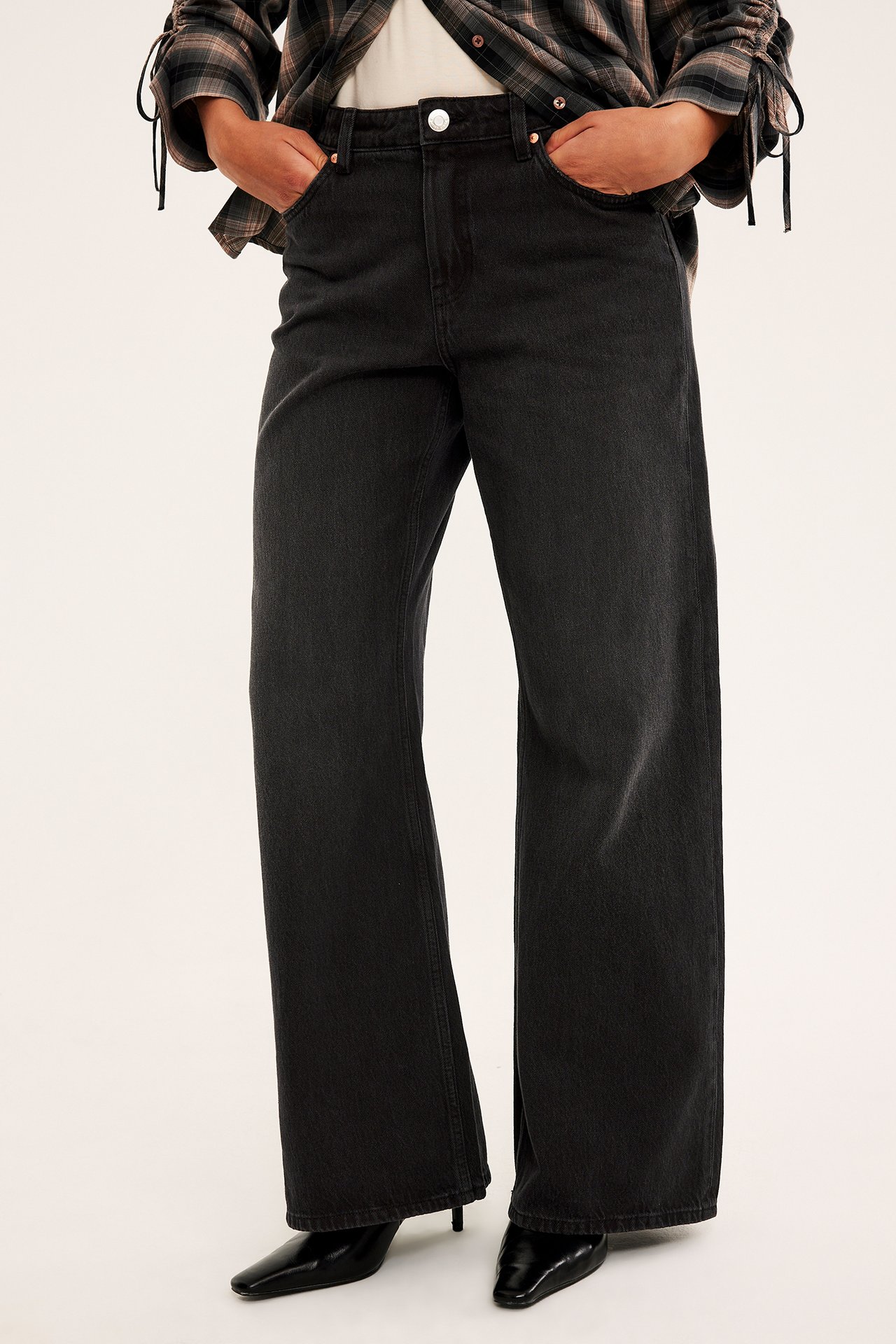Front av kvinnelig modell i mørkegrå vide denimjeans, rutete skjorte og svarte hælstøvler.