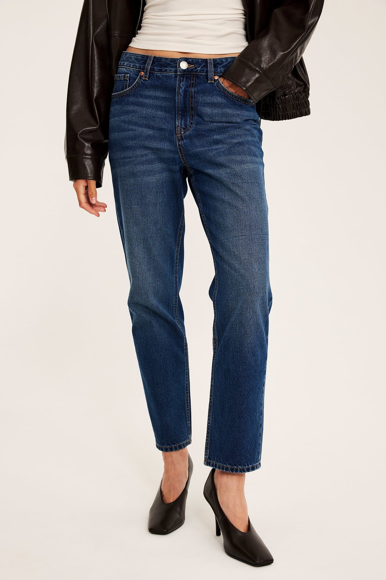 Frontvy av en kvinna i blå raka jeans, vit linne, svart läderjacka och svarta klackskor.