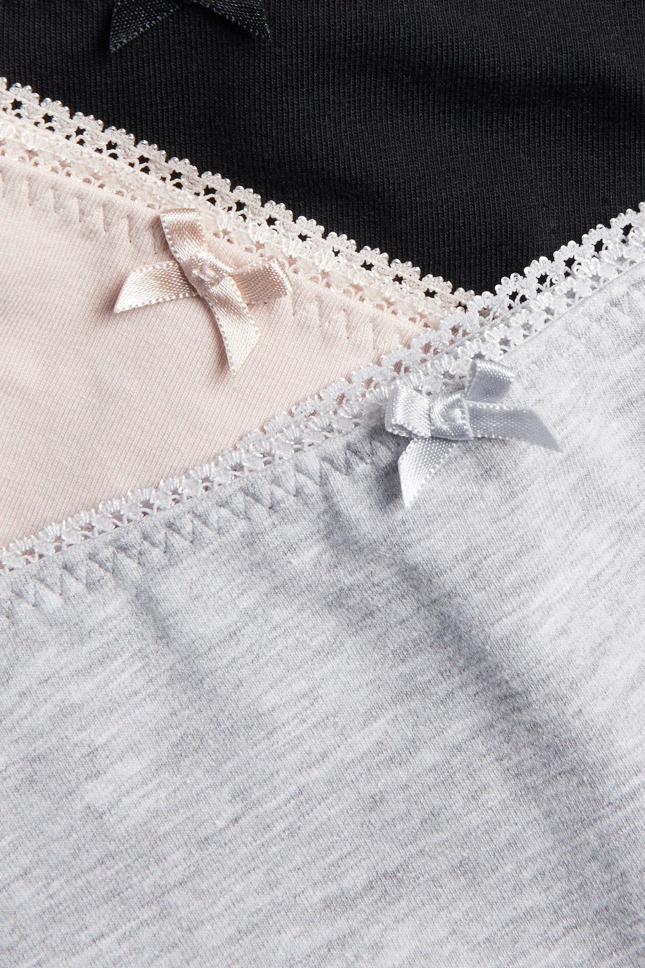 Nærbilde av tre stablede bomullsstringtruser: svart, lys rosa og lys grå med picotkanter og små sløyfer.