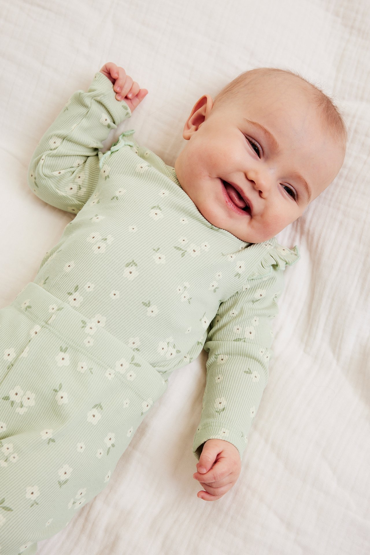 Ylhäältä kuvattu hymyilevä vauva yllään vaaleanvihreä ribbineulebody valkoisella kukkakuviolla ja olkarypyillä.