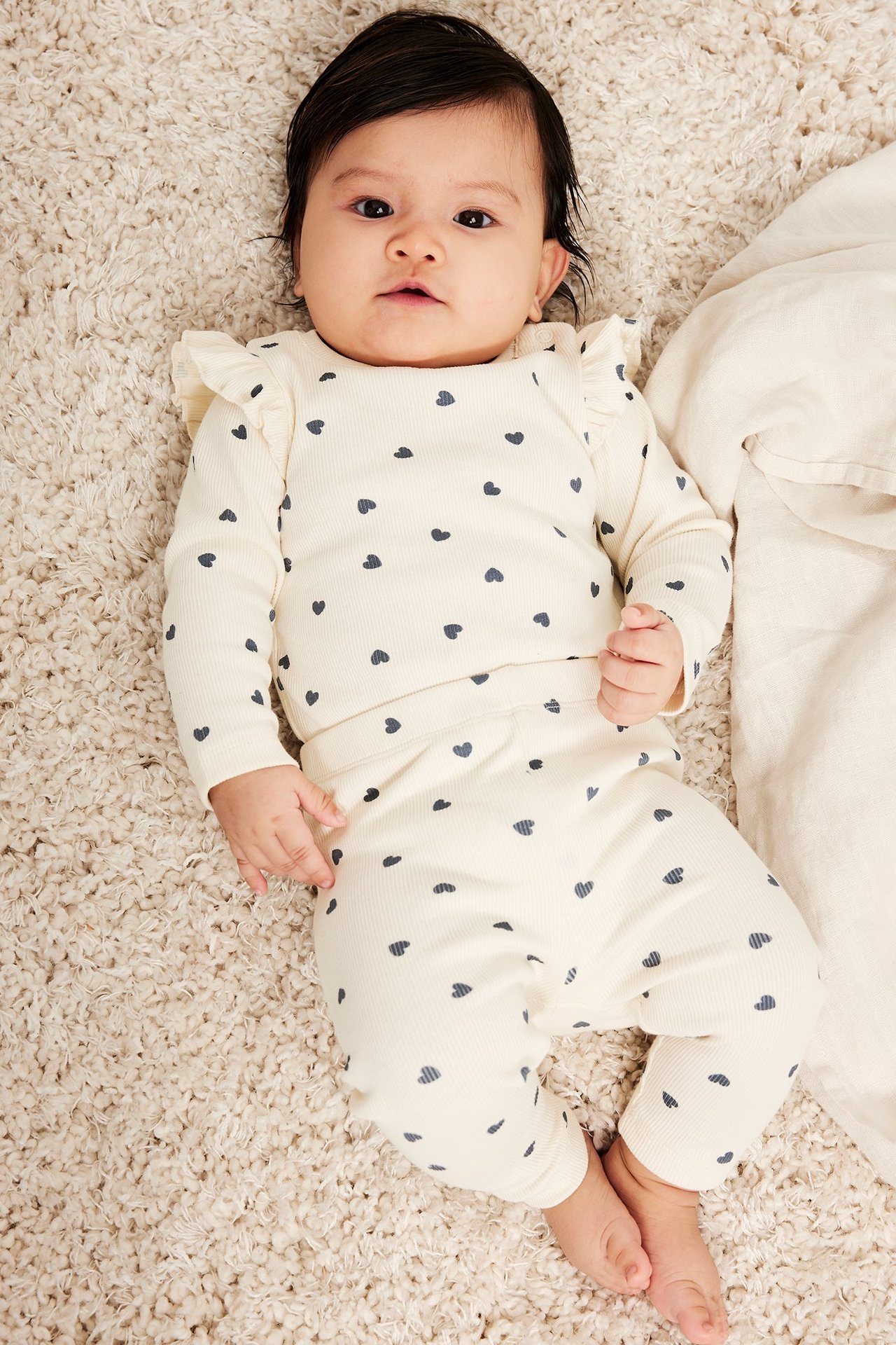 Toppvy av en baby i en offwhite ribbad långärmad body med hjärtmönster och axelvolanger, matchande byxor.