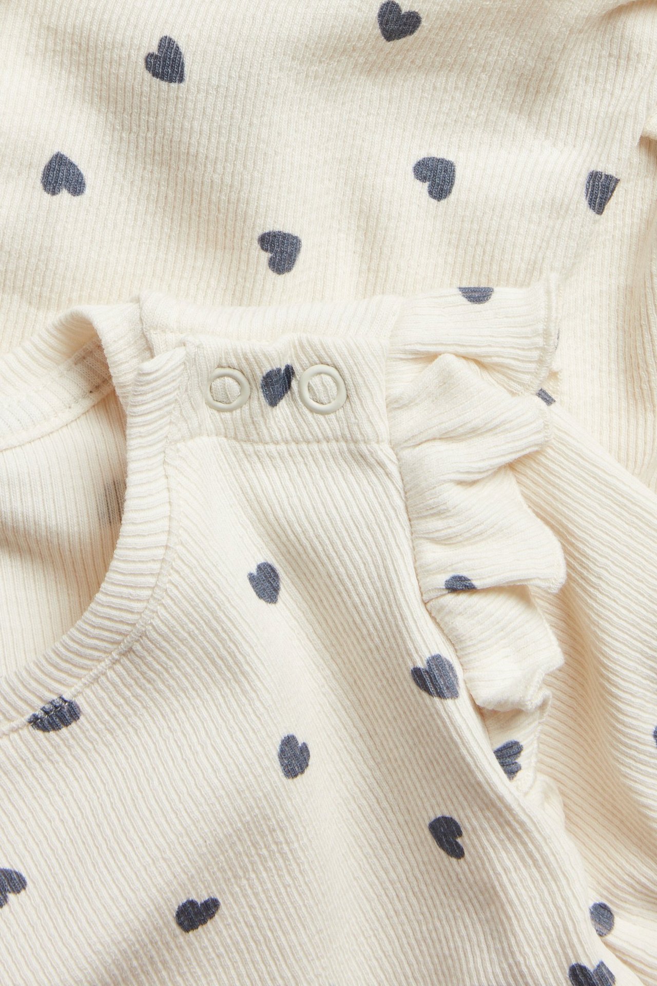 Lähikuva luonnonvalkoisesta ribatusta vauvanbodysta, jossa on tumma sydänkuvio, olkapään röyhelöt ja painonapit.