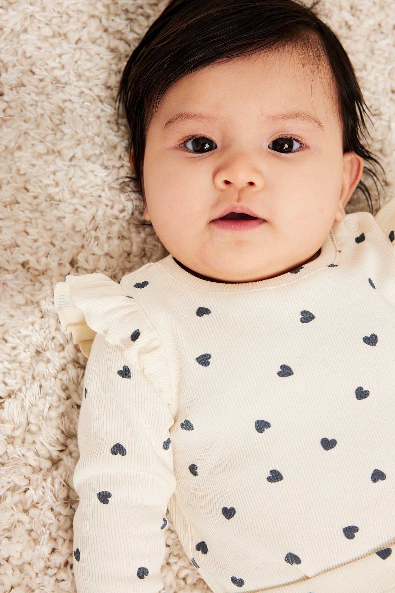 Närbild framifrån av en baby i offwhite ribbad body med mörkt hjärtmönster och volanger.
